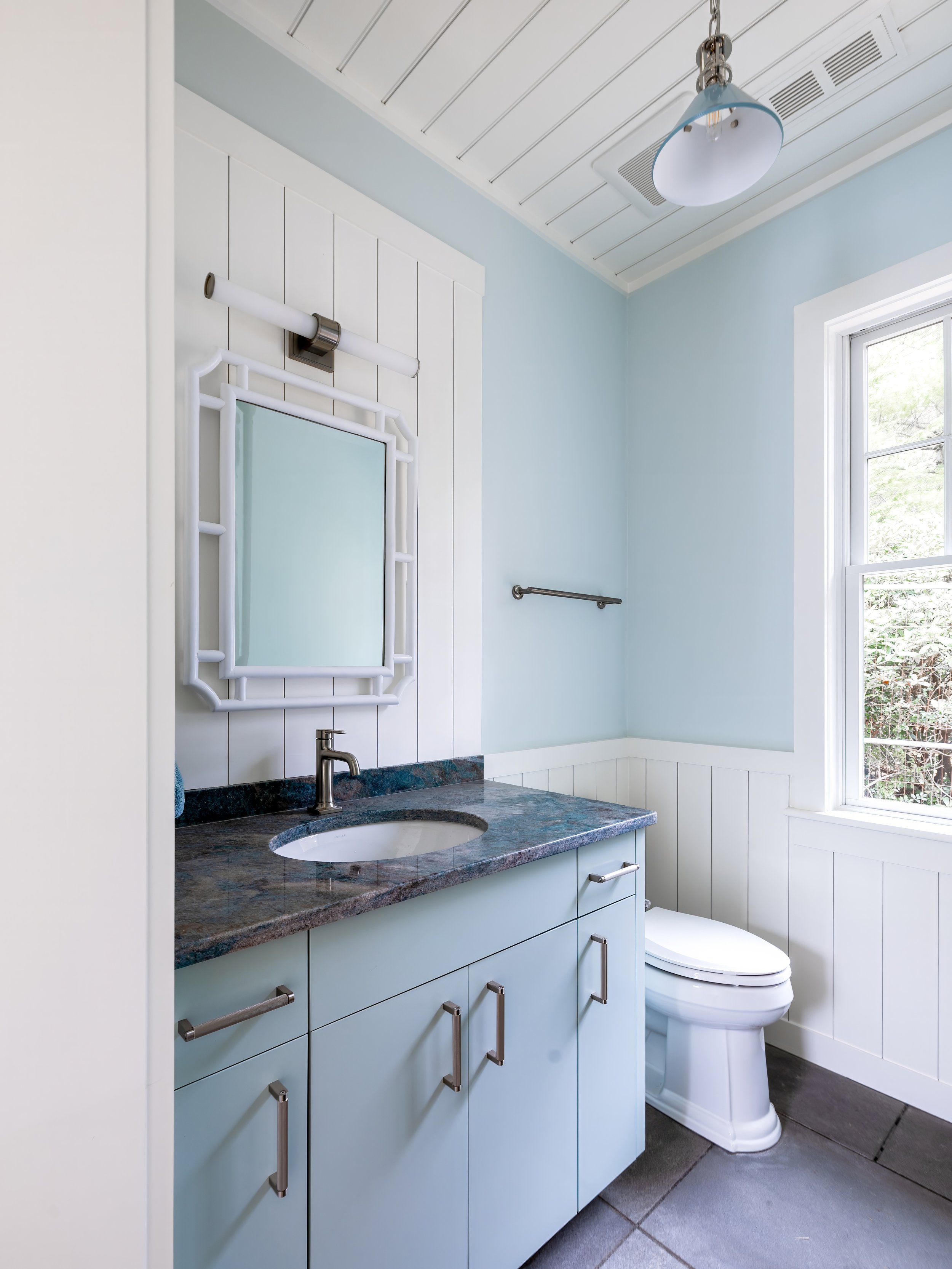 Cape Cod bathroom with blue vanity, stone countertop, white paneling, and natural light. 