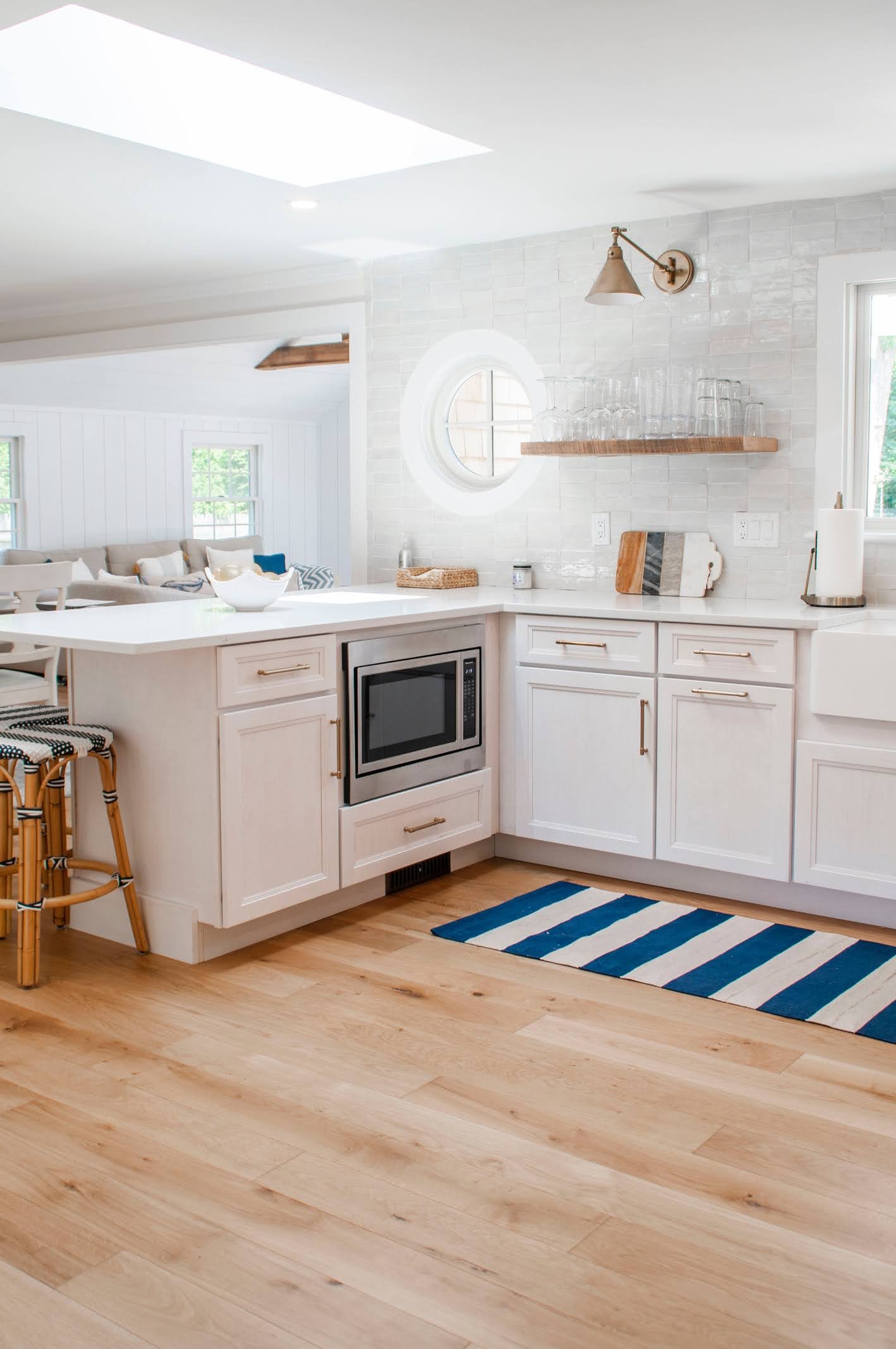 an airy custom coastal home kitchen showcases white cabinetry, wood floors, round window, and classic Cape Cod charm. 