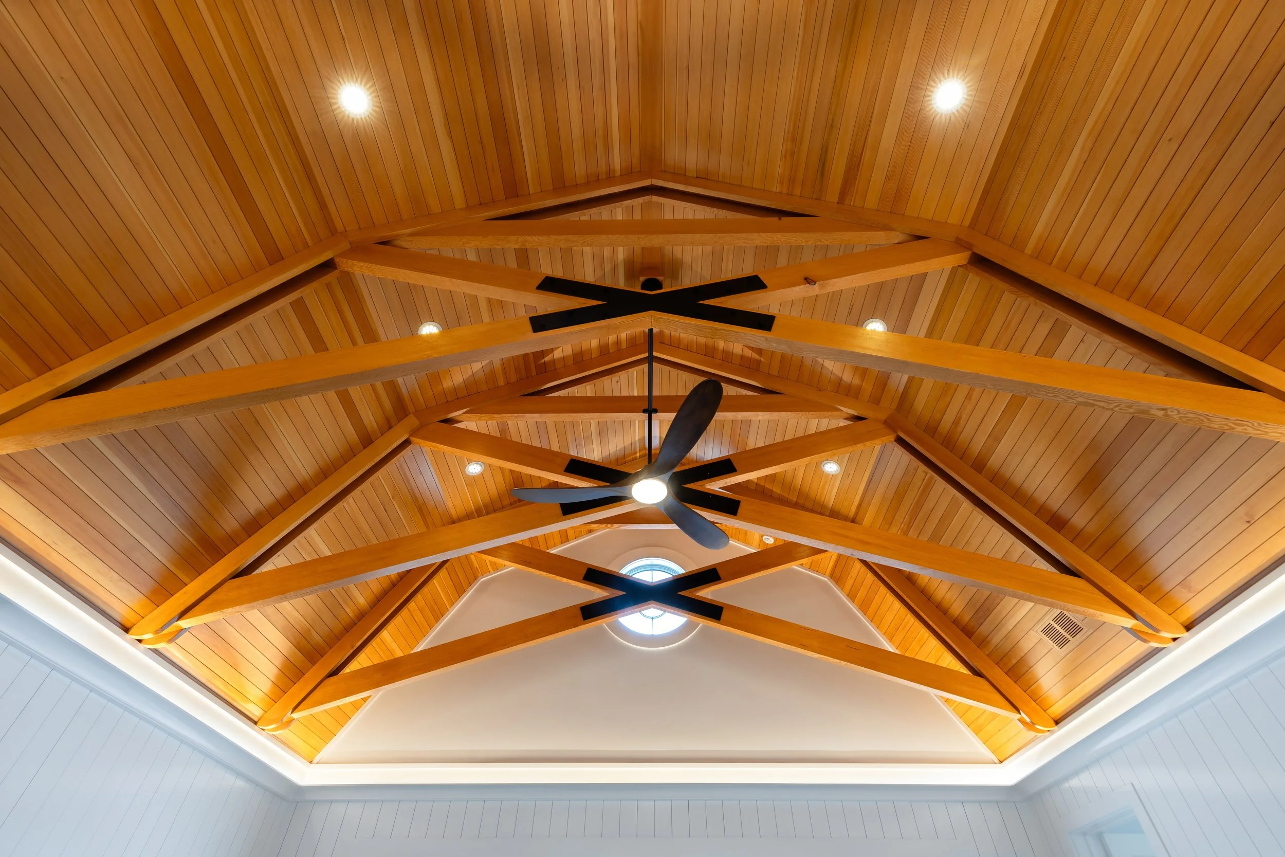 High ceilings with exposed wood trusses and fan in a shingle-style Cape Cod pool house.