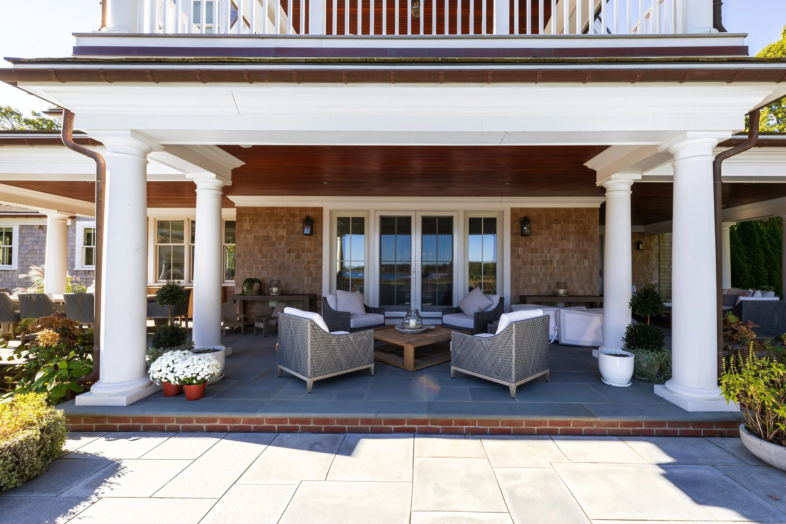 Covered porch with white columns, wicker lounge seating, wood ceiling, and waterfront views beyond glass doors.
