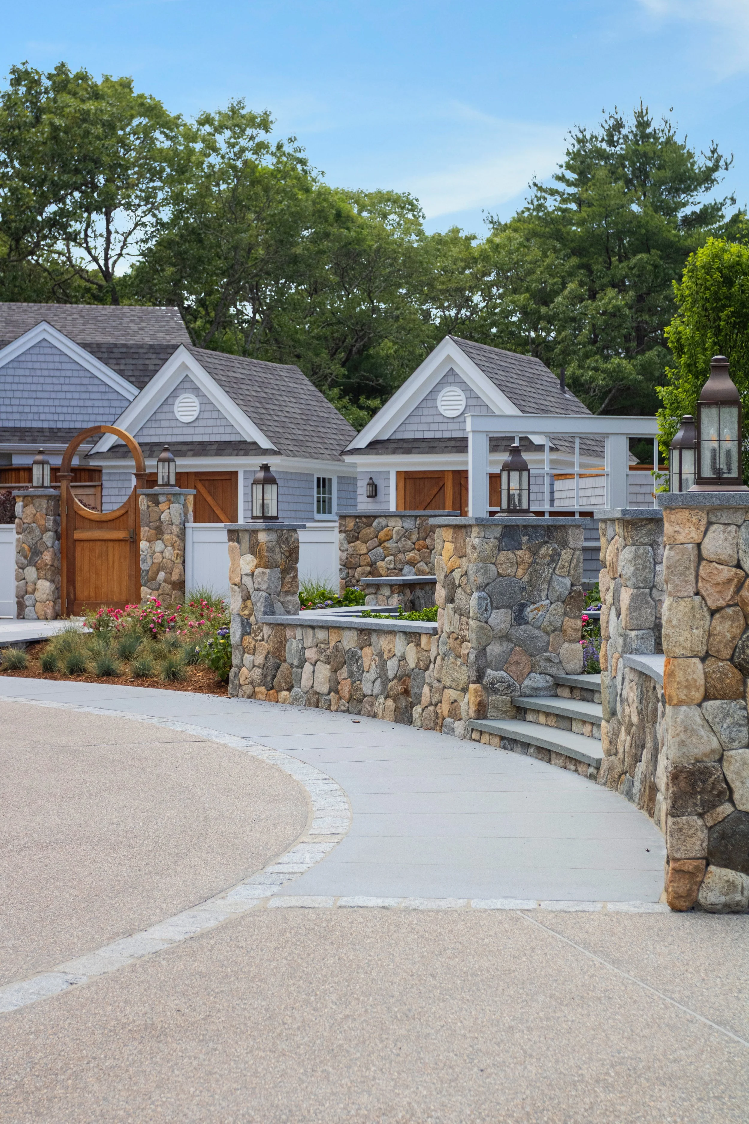 Natural stone walls, custom cedar fencing, and shingle-style structures define this professionally designed Cape Cod landscape and outdoor entertaining area.