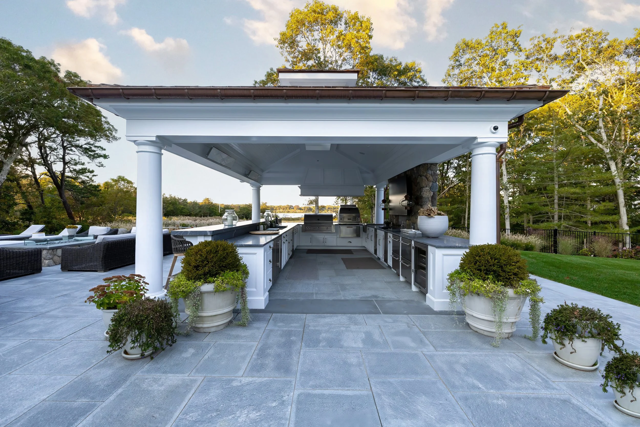 Covered outdoor kitchen with white columns, potted greenery, stone patio, and open Cape Cod views.