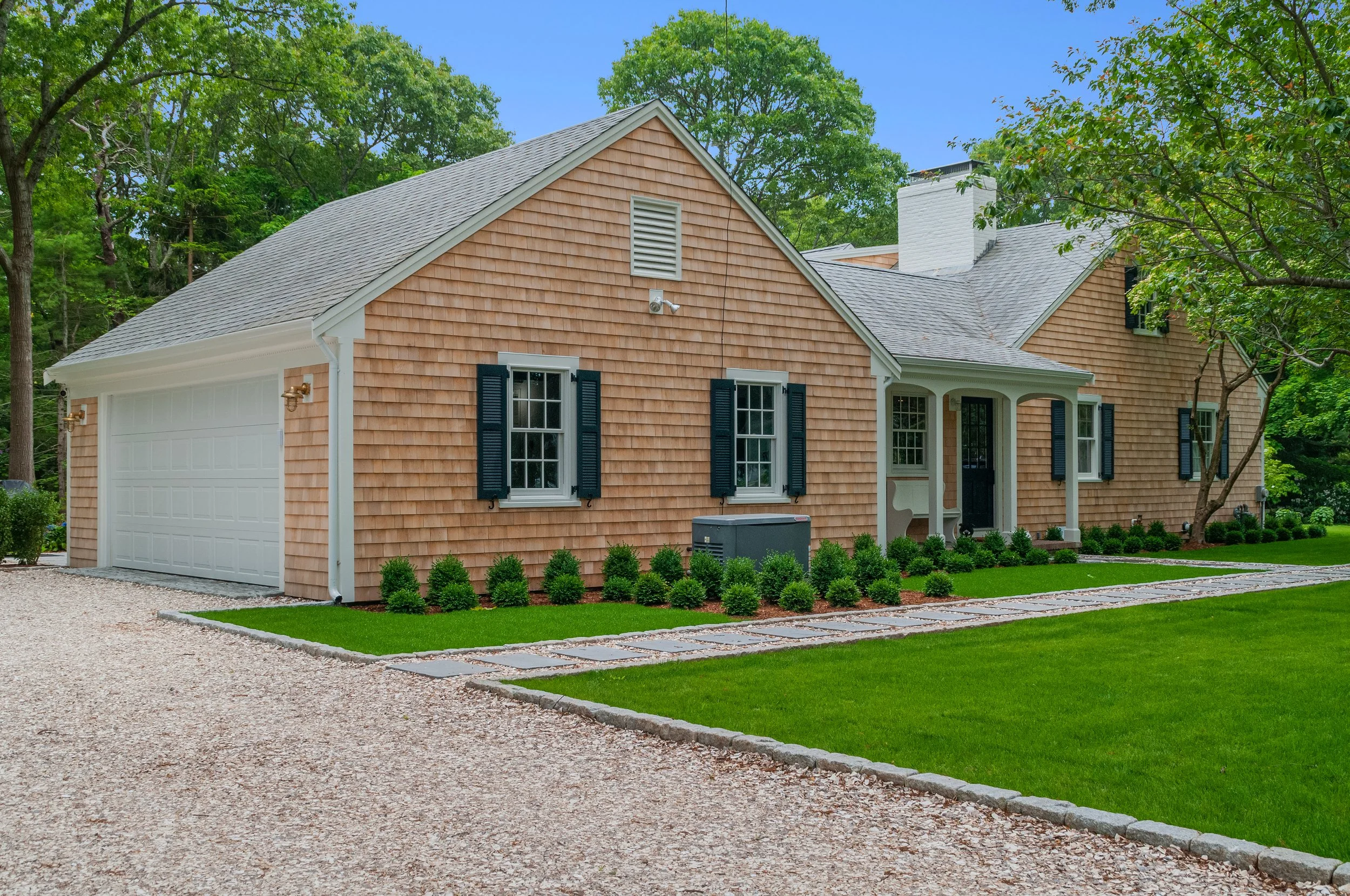 Attached garage, black shutters, gravel drive, and coastal landscaping define this custom Cape Cod home.