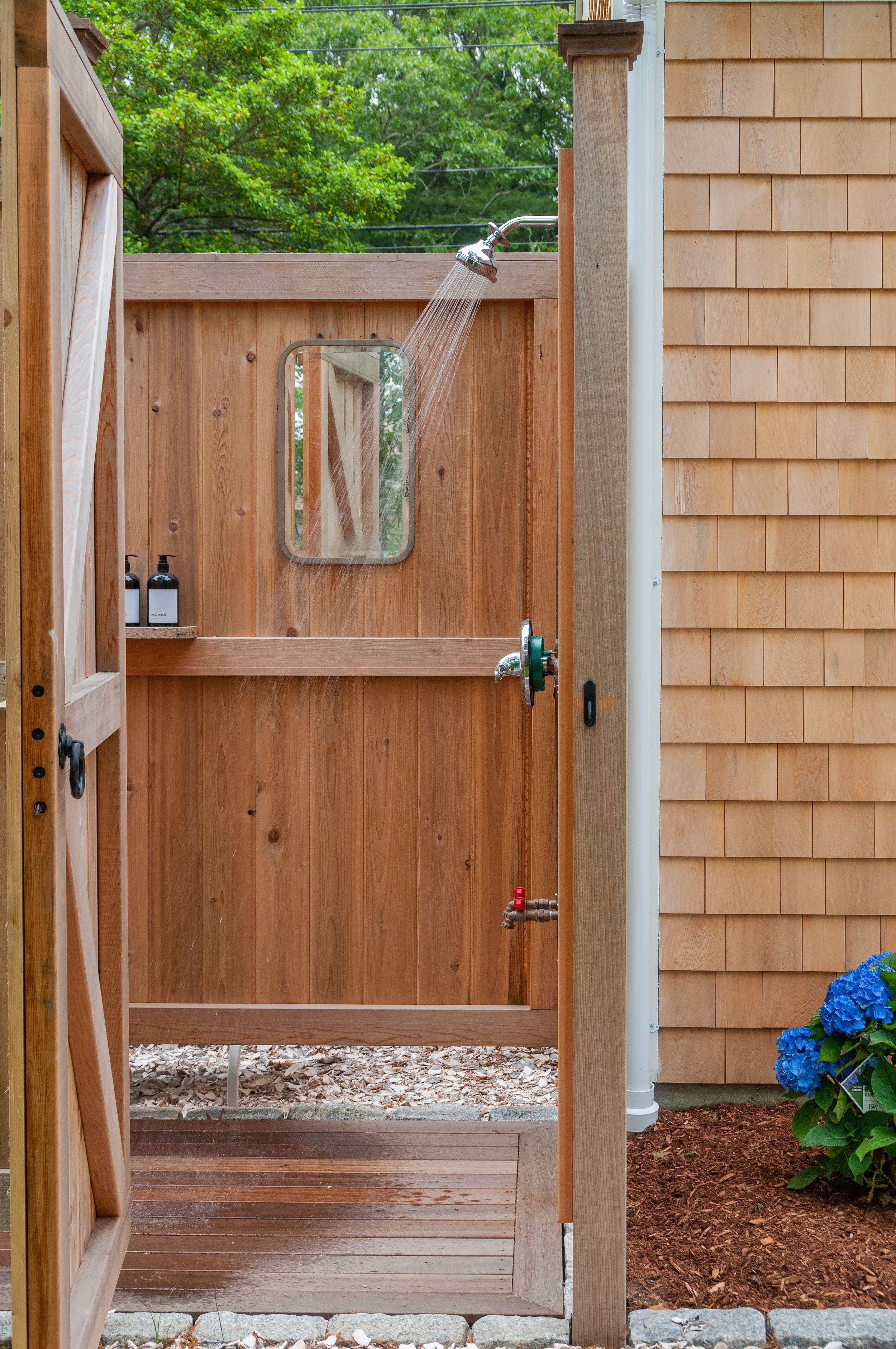 Outdoor shower with cedar enclosure, shingled Cape Cod siding, wood decking, and blue hydrangeas.