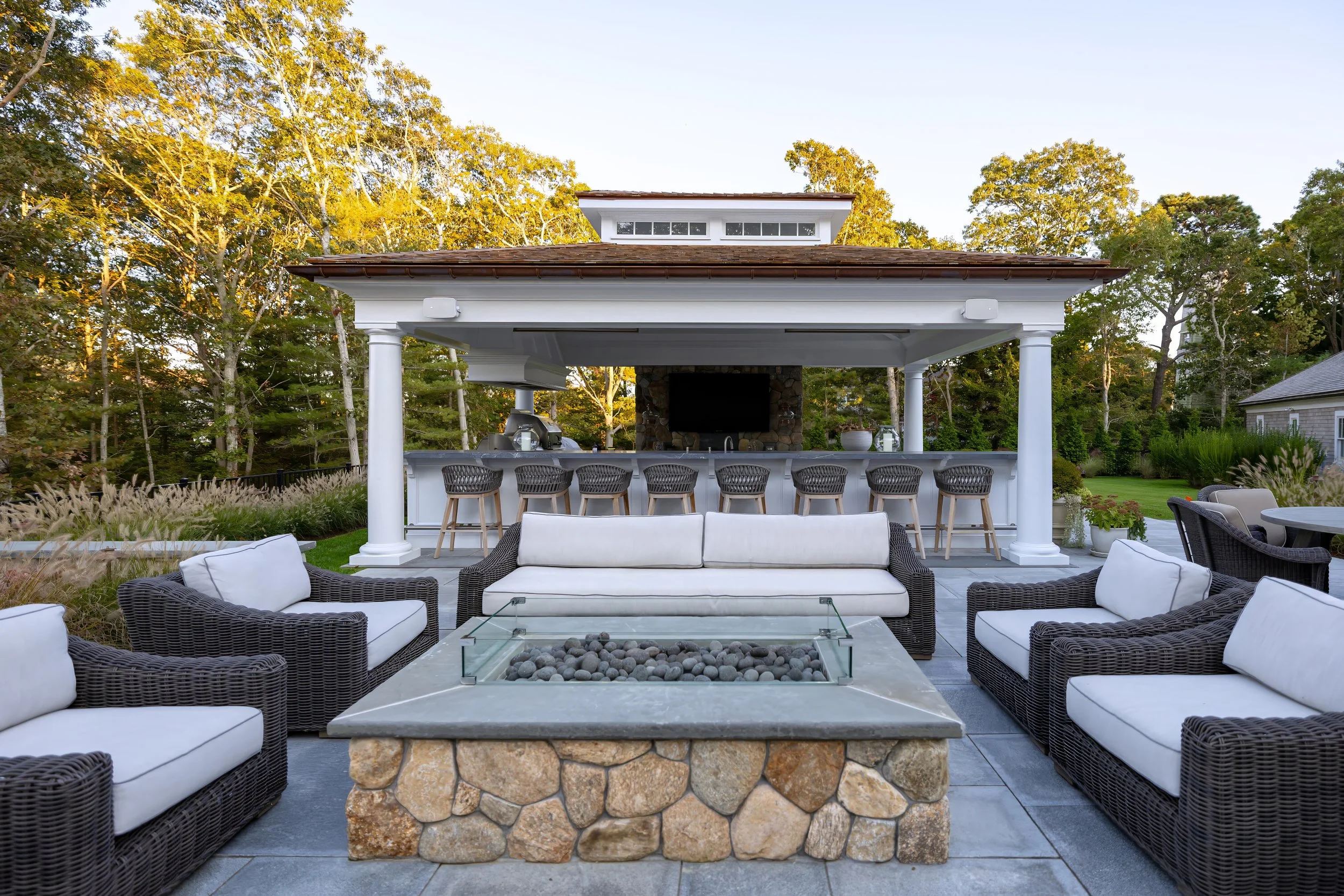 Covered outdoor living area with stone fire feature, wicker seating, bar stools, and wooded Cape Cod views.
