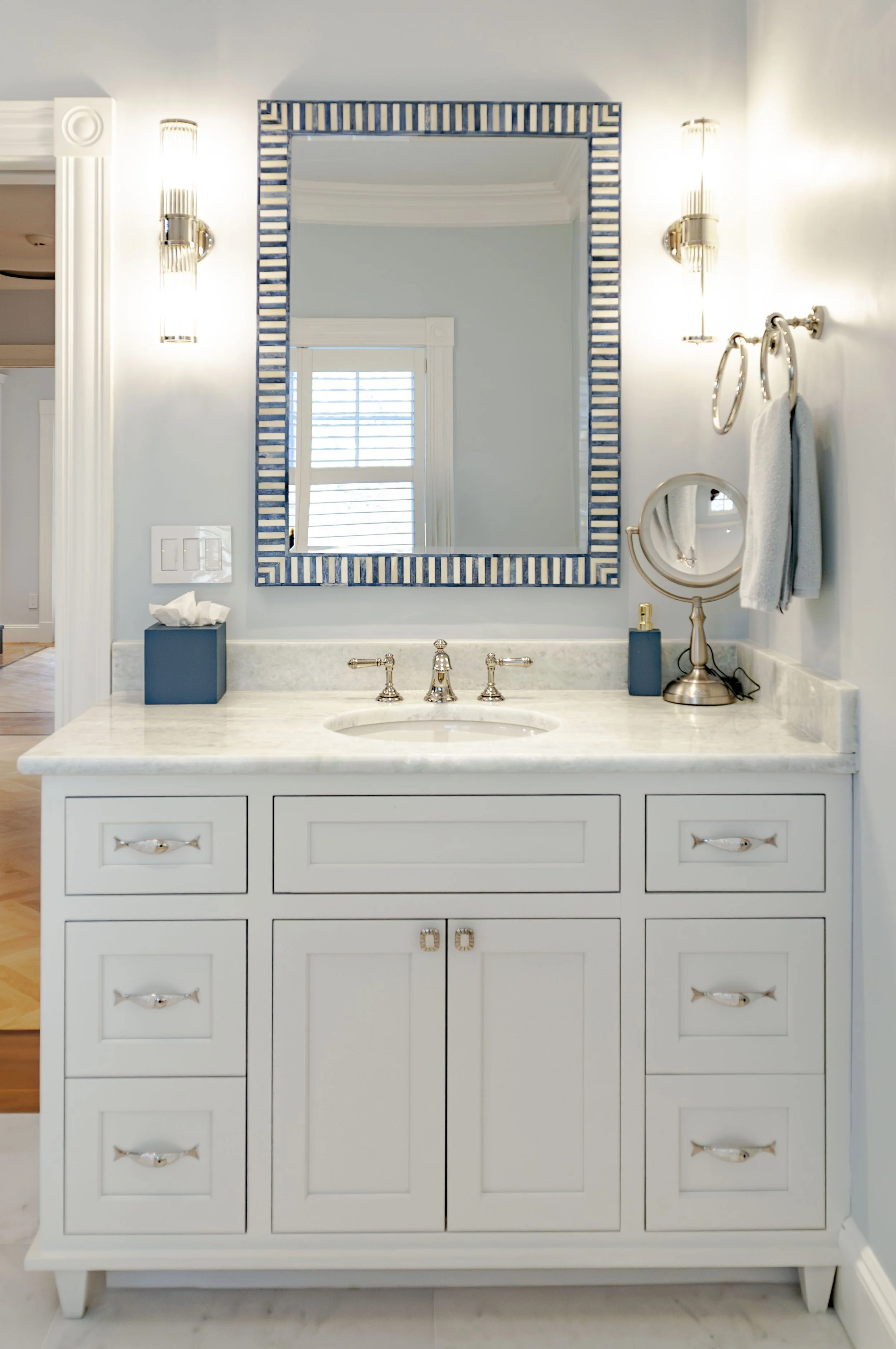 Bathroom vanity with marble counter, blue-trimmed mirror, polished sconces, and custom cabinetry.