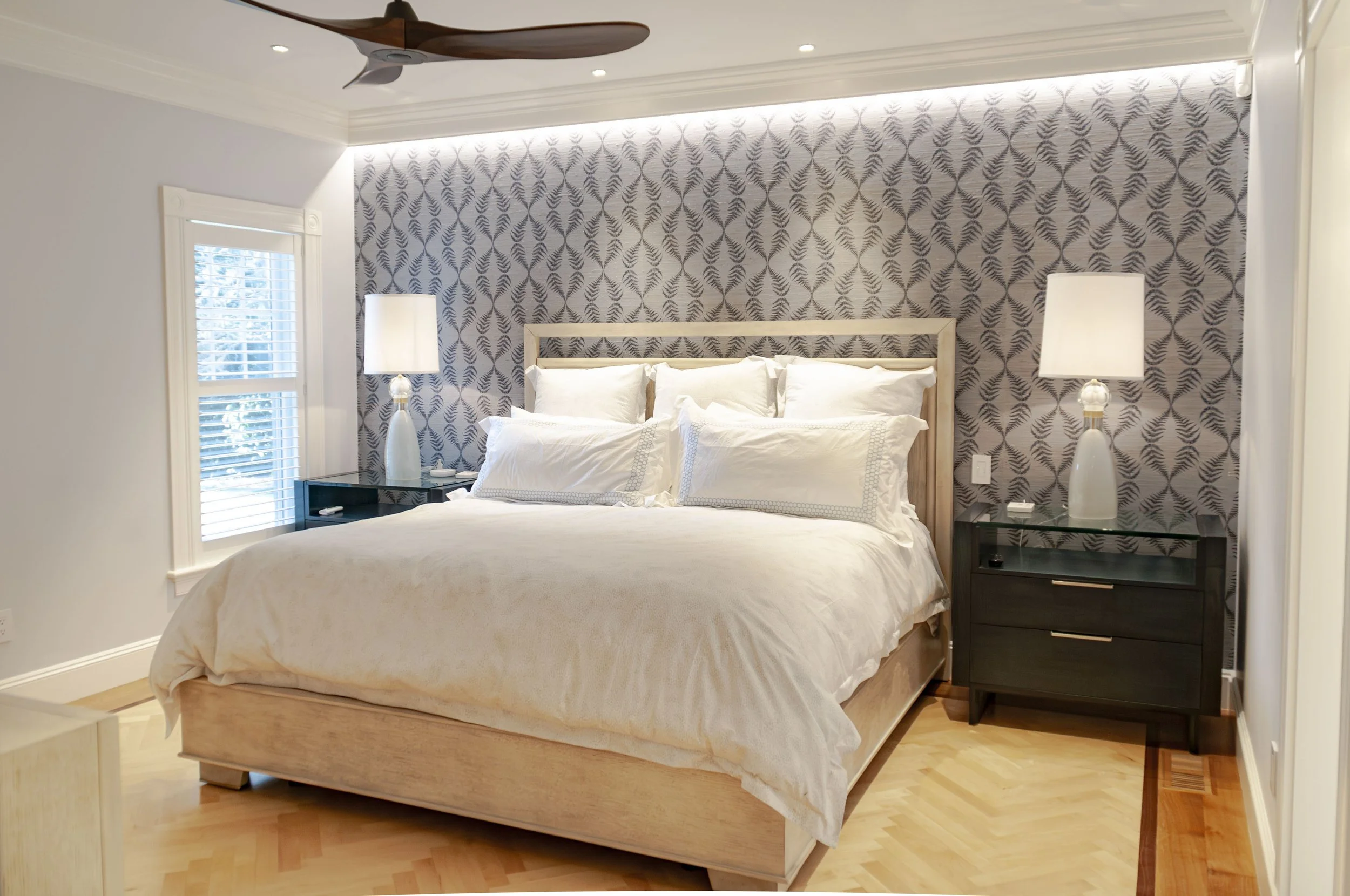 Cape Cod bedroom with patterned accent wall, white bedding, wood ceiling fan, and herringbone flooring.