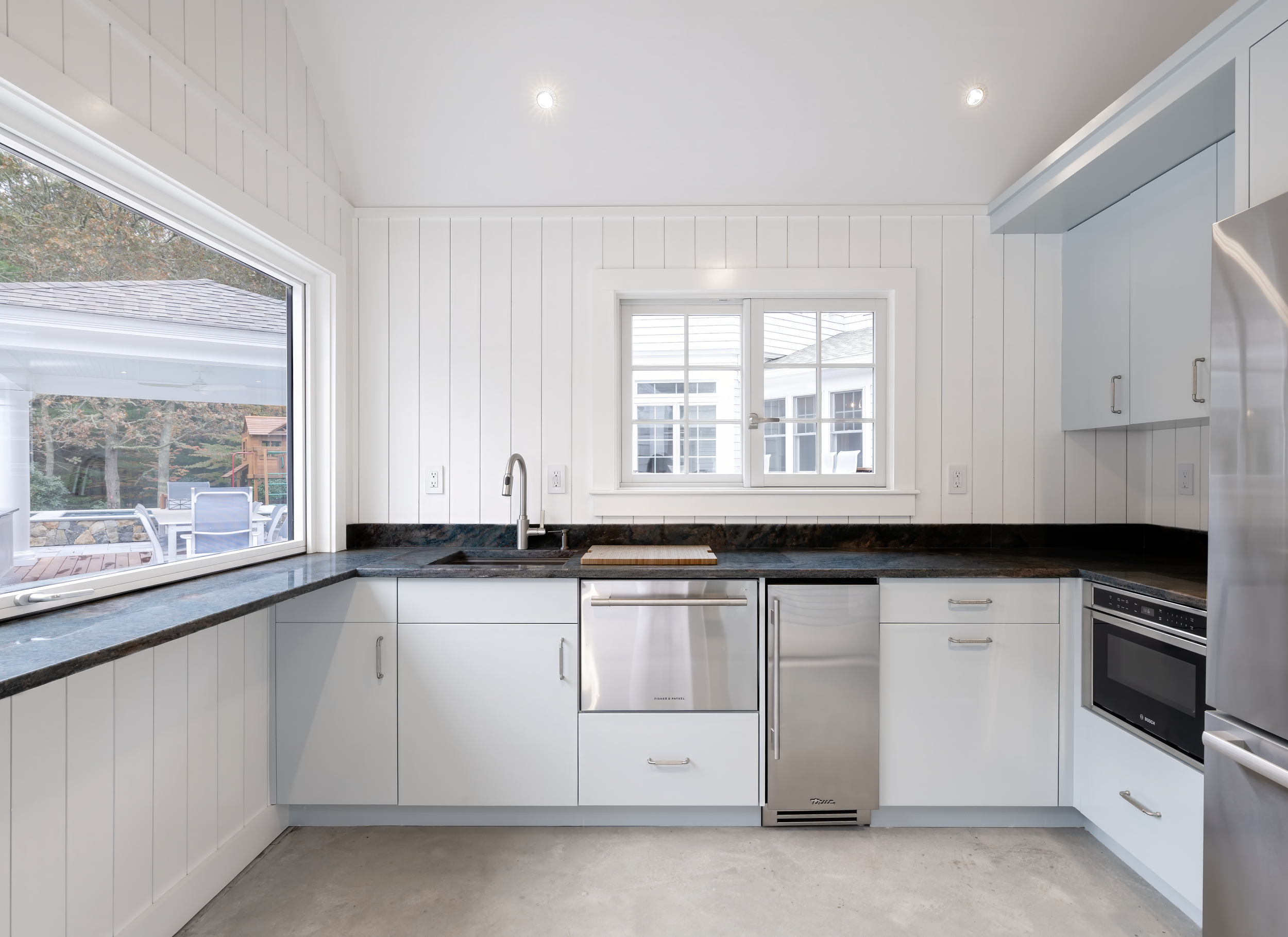 Bright pool house kitchenette with white paneling, black countertops, stainless appliances, and poolside views.
