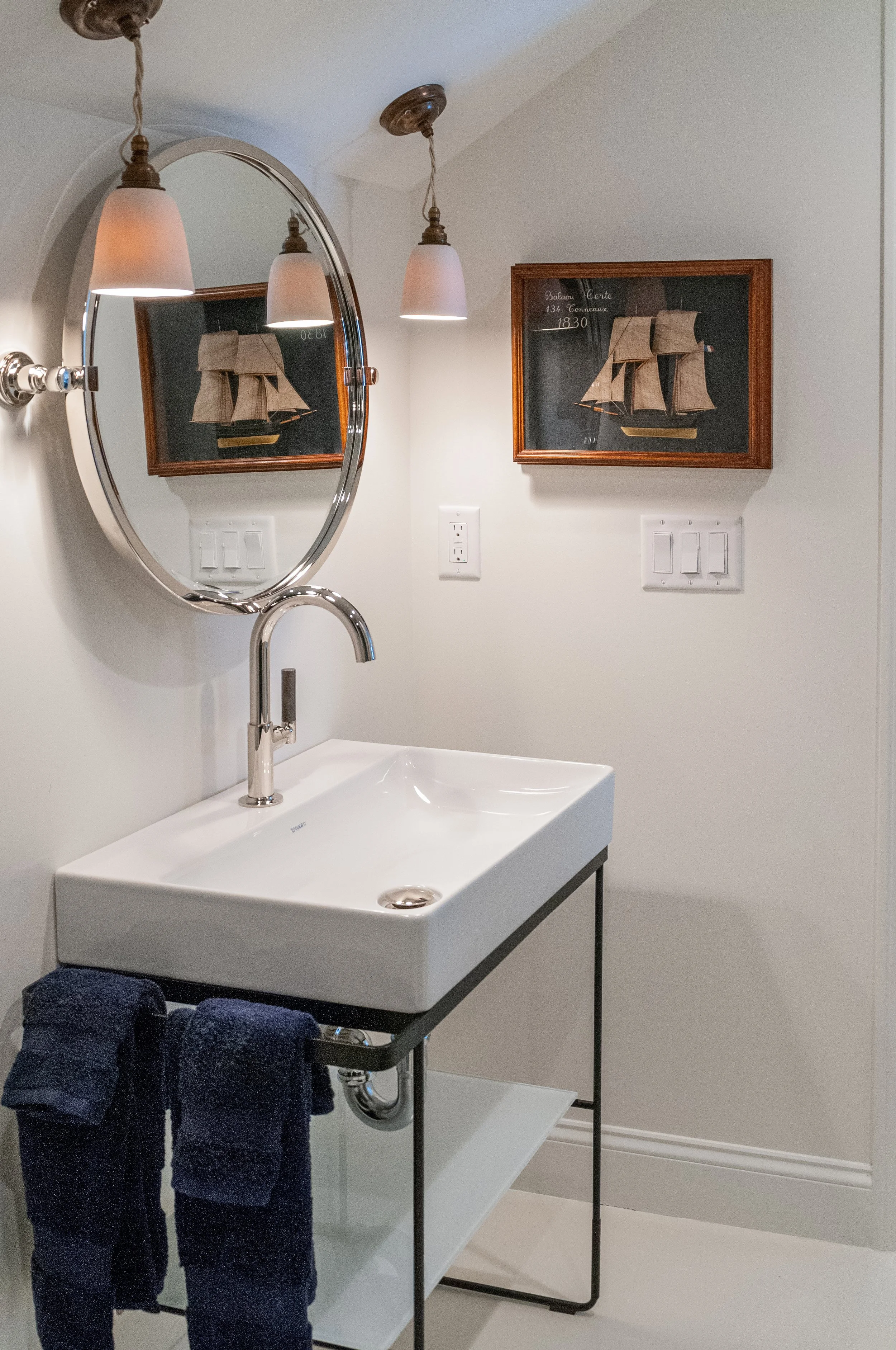 Custom Cape Cod powder room with wall-mounted sink, round mirror, pendant lights, and nautical artwork.
