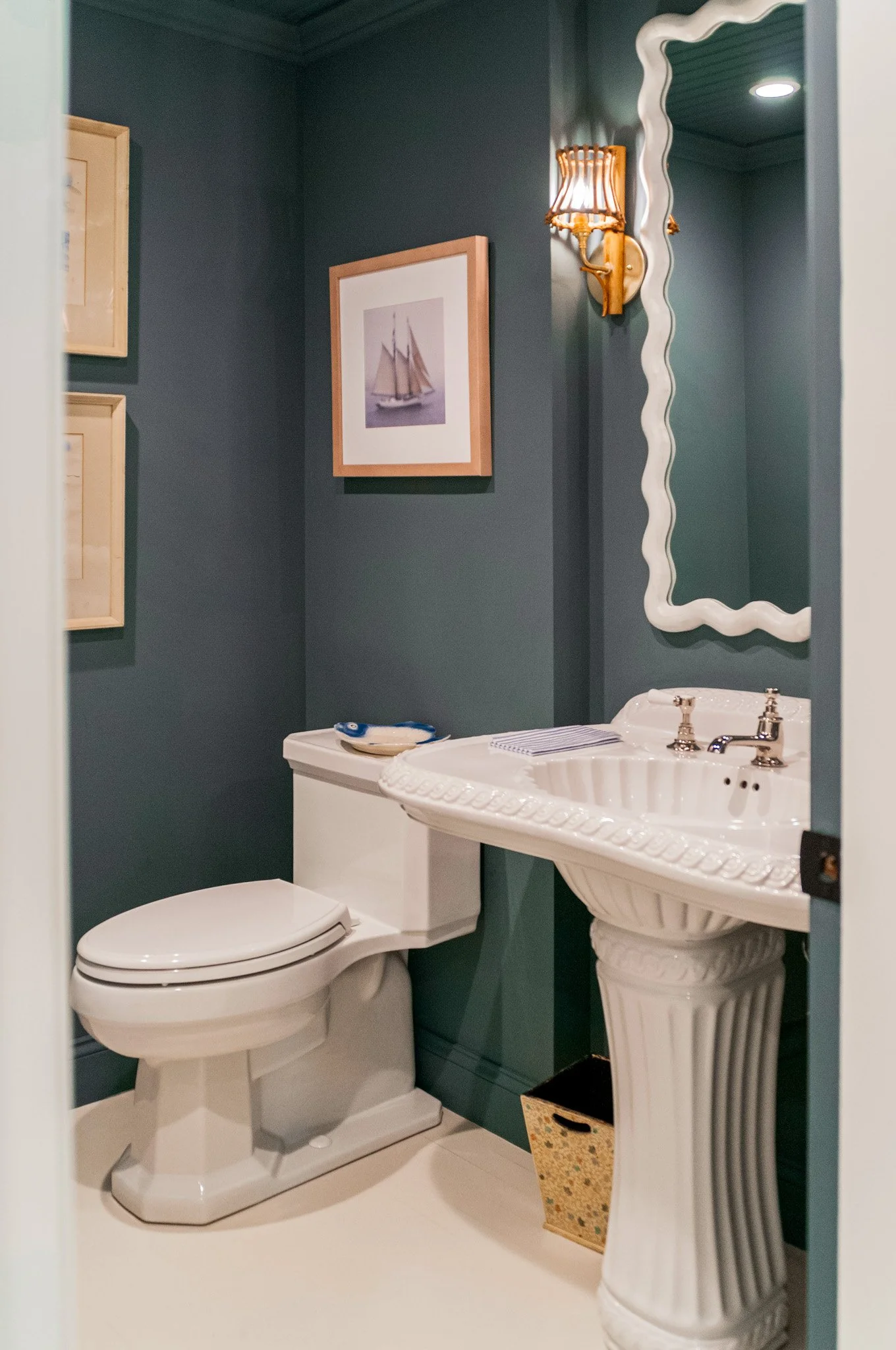 Dark painted powder room with pedestal sink, sculptural mirror, and coastal bathroom design details.