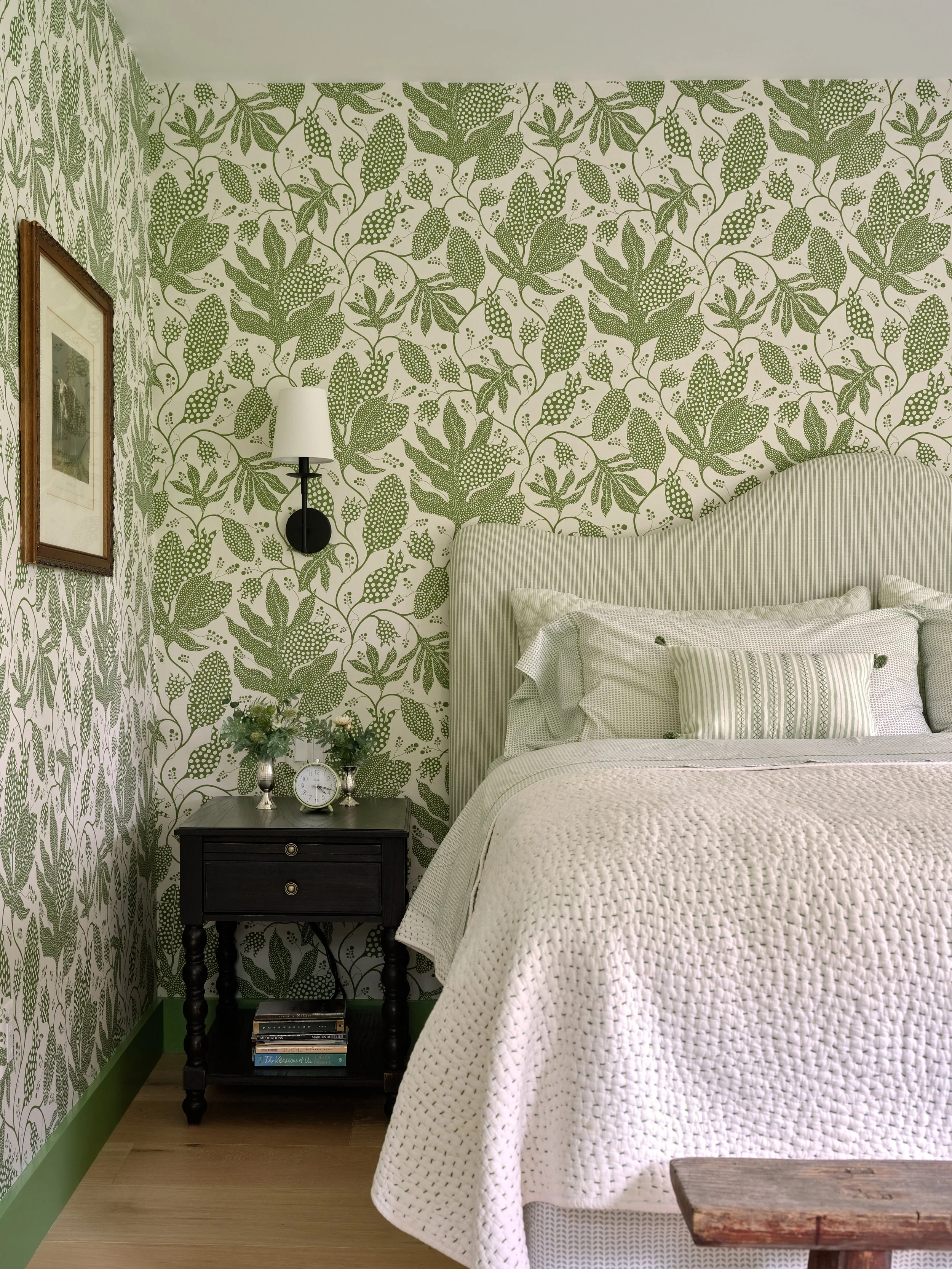 Bedroom with patterned green wallpaper, upholstered headboard, black nightstand, and soft layered bedding in Cape Cod style.