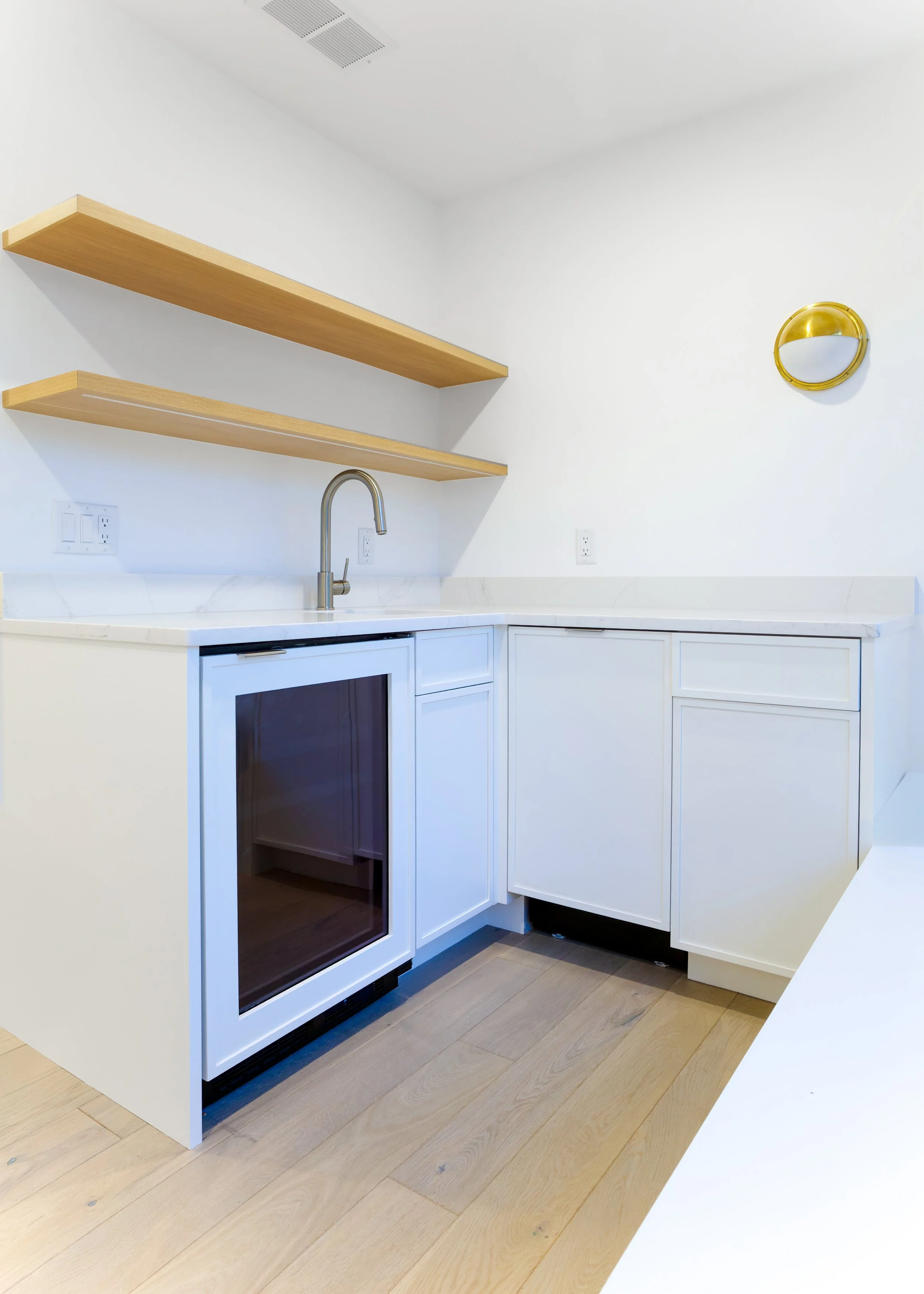 White kitchenette with wood floating shelves, sink, beverage fridge, and bright coastal guest suite design.