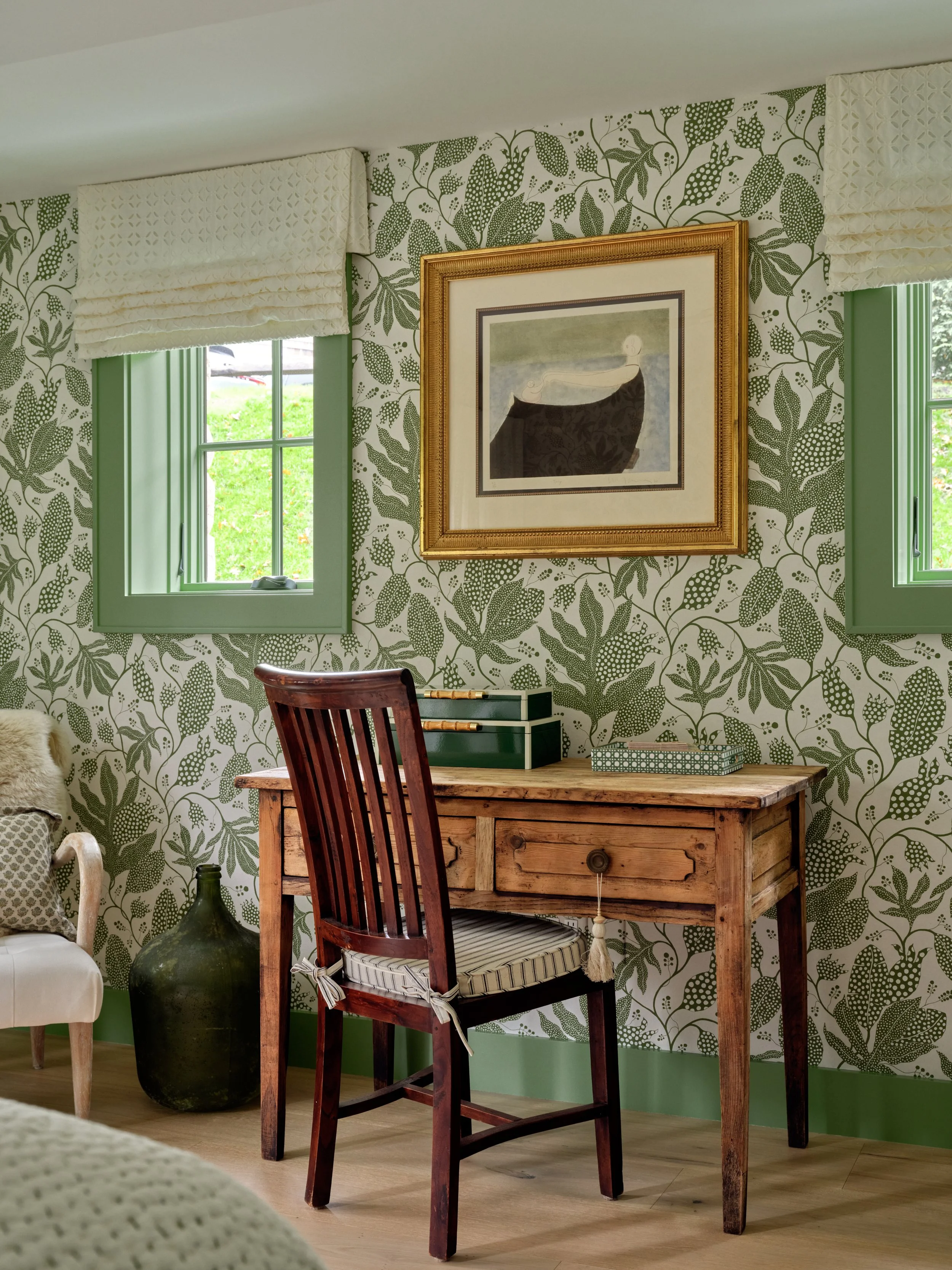Bedroom writing nook with botanical wallpaper, green-trimmed windows, framed art, and rustic wood desk.