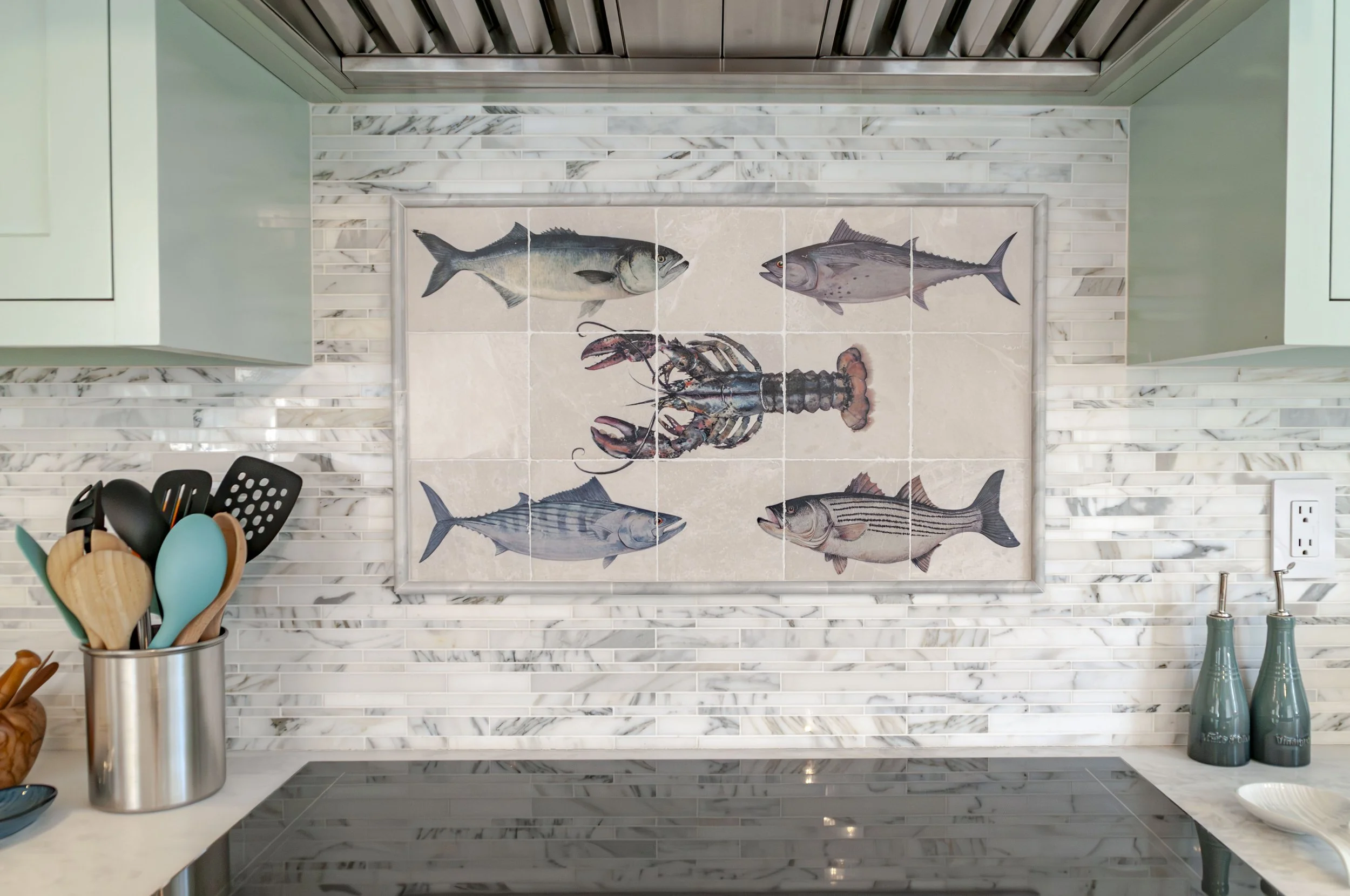 Cape Cod kitchen detail with marble backsplash, cooktop, range hood, and coastal fish tile artwork.