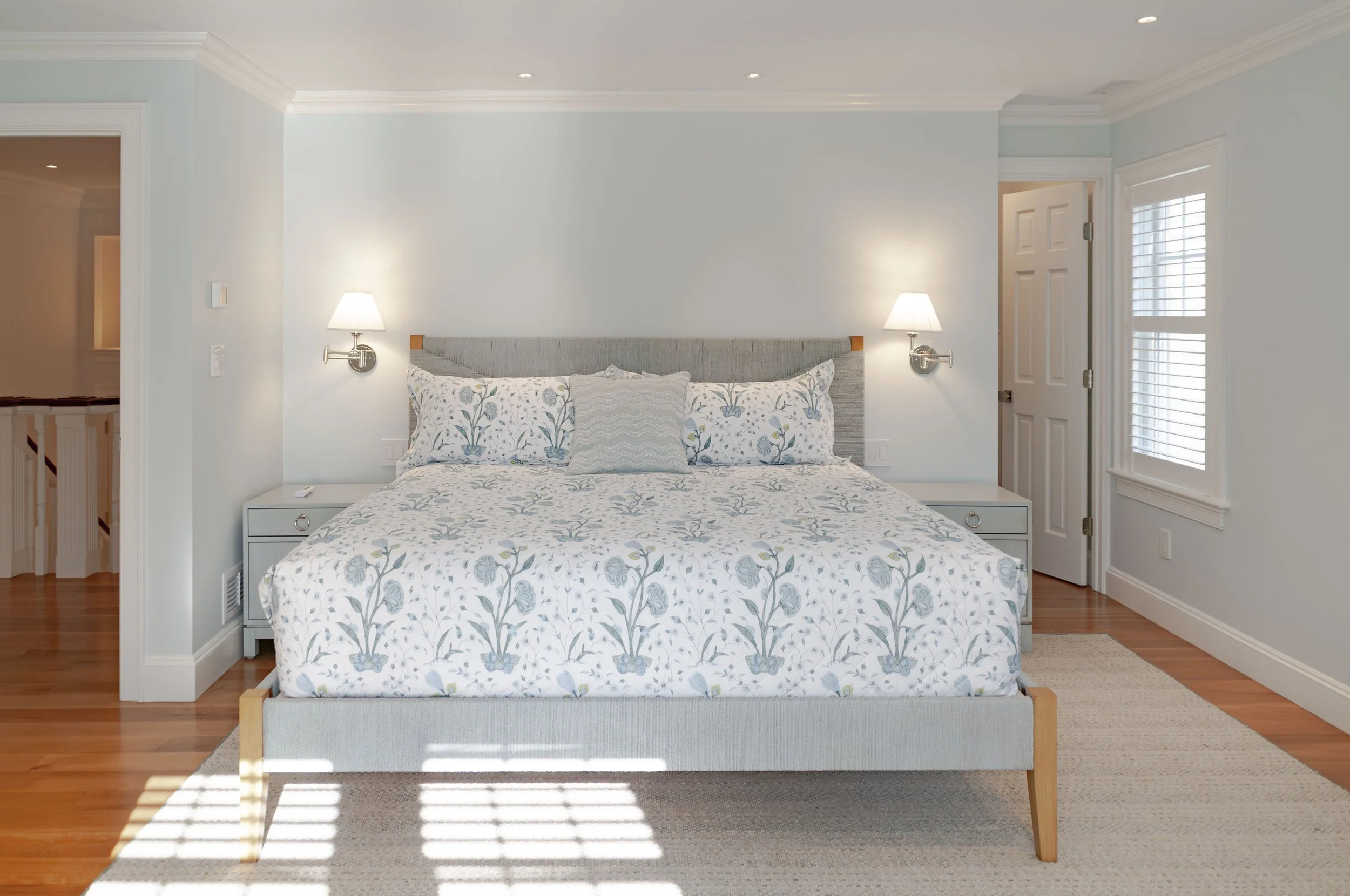 Cape Cod bedroom with soft blue walls, floral bedding, shuttered window, and custom coastal home finishes.