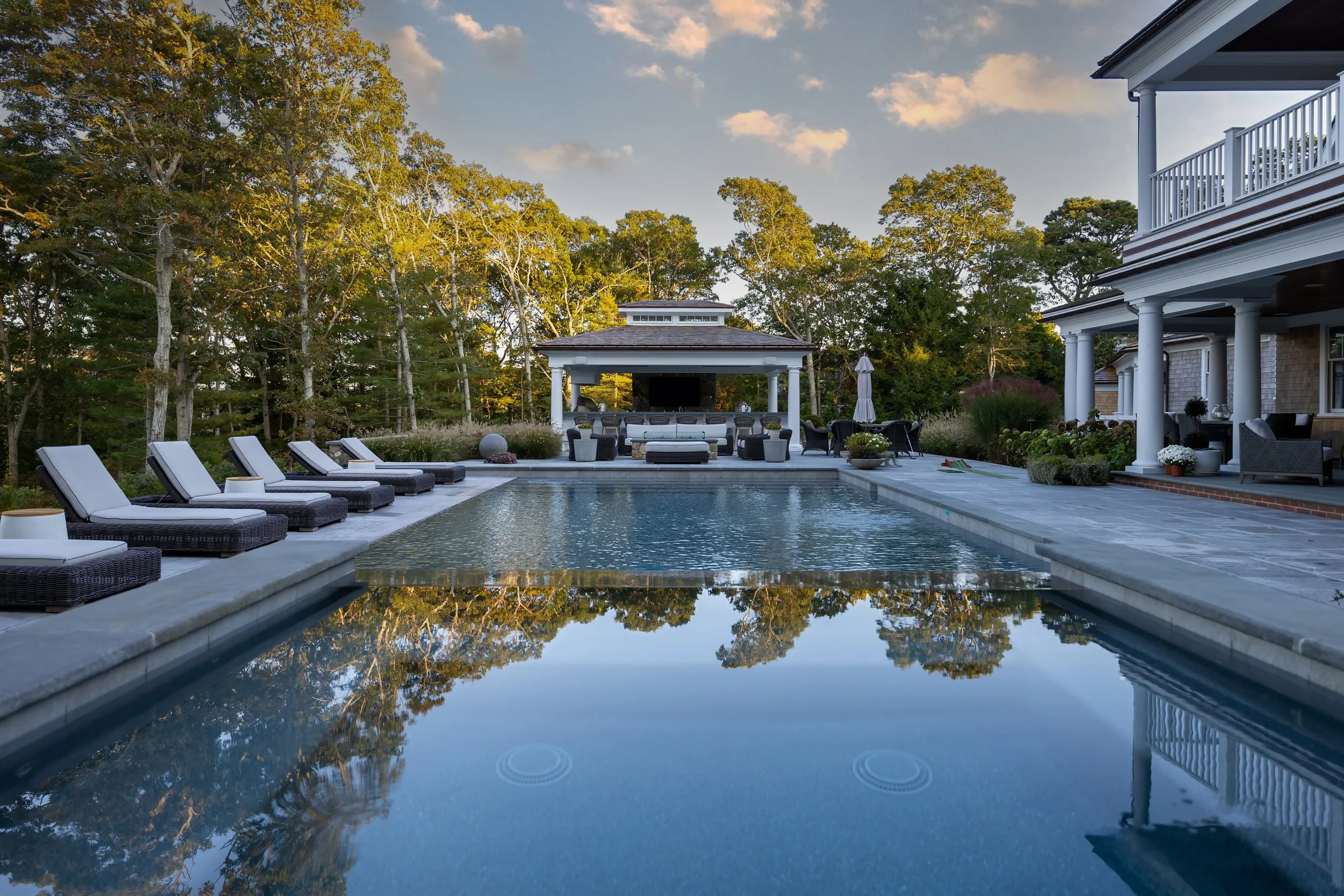 Luxury poolside lounge at a custom coastal home with  covered cabana, white columns, and wooded Cape Cod backdrop.