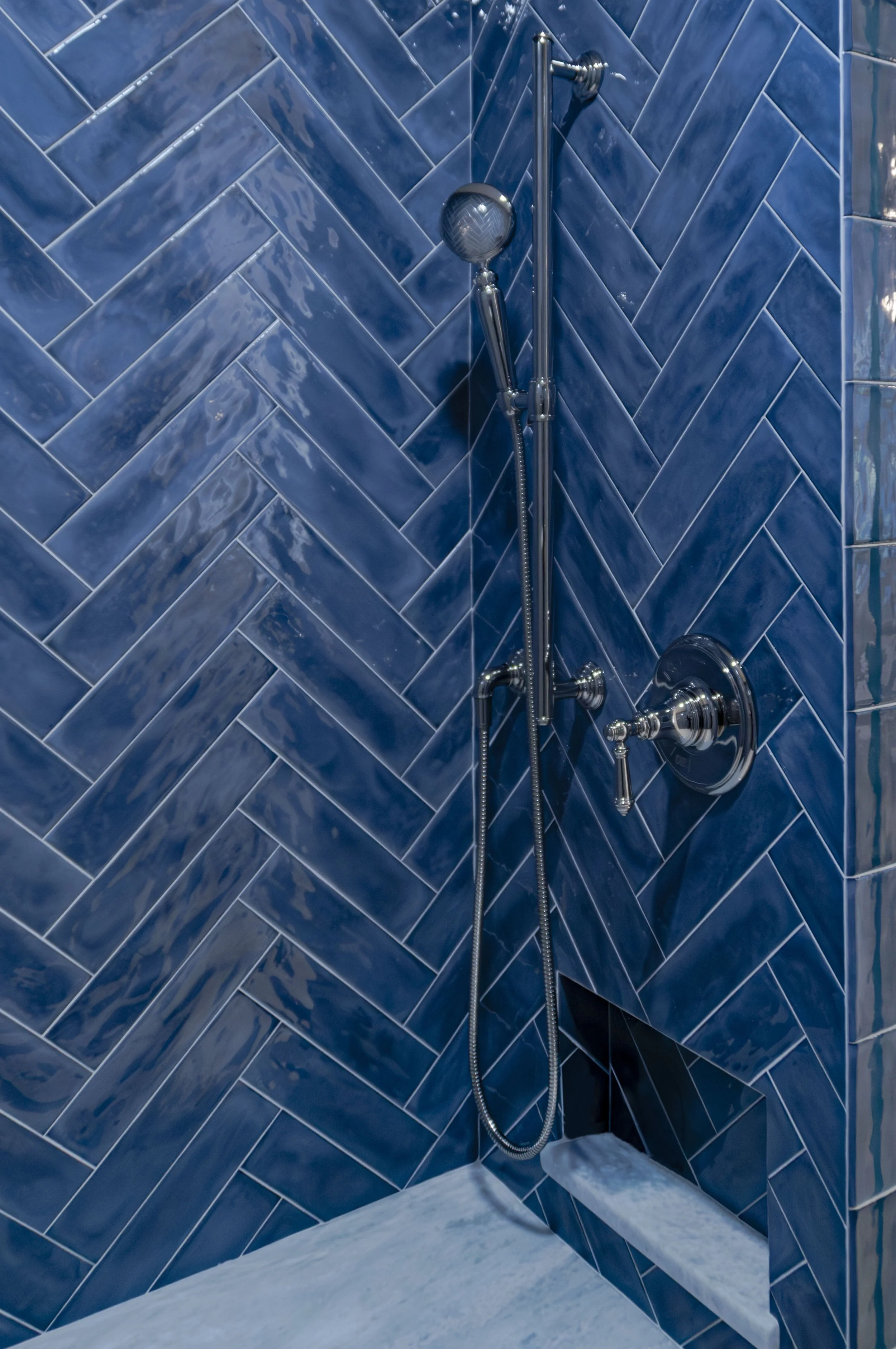 Cape Cod shower detail with glossy blue herringbone tile, handheld fixture, and marble bench.
