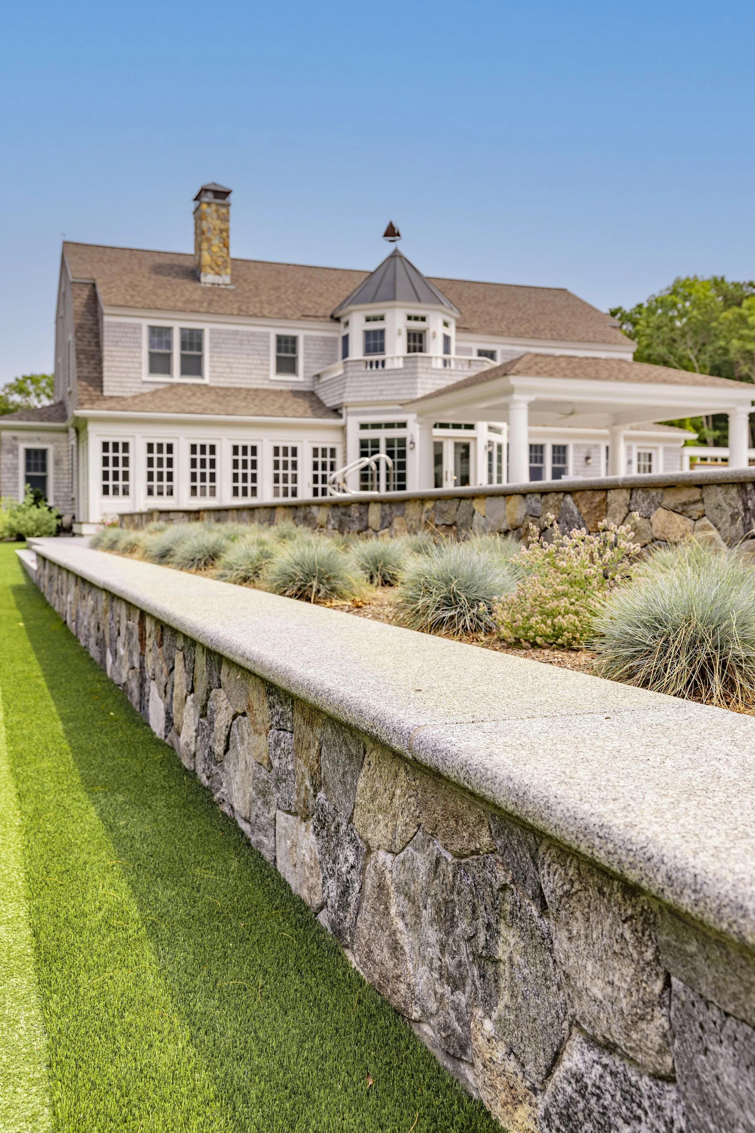 Stone retaining wall and embellishing grasses frame a shingle-style cape cod home with covered teracce