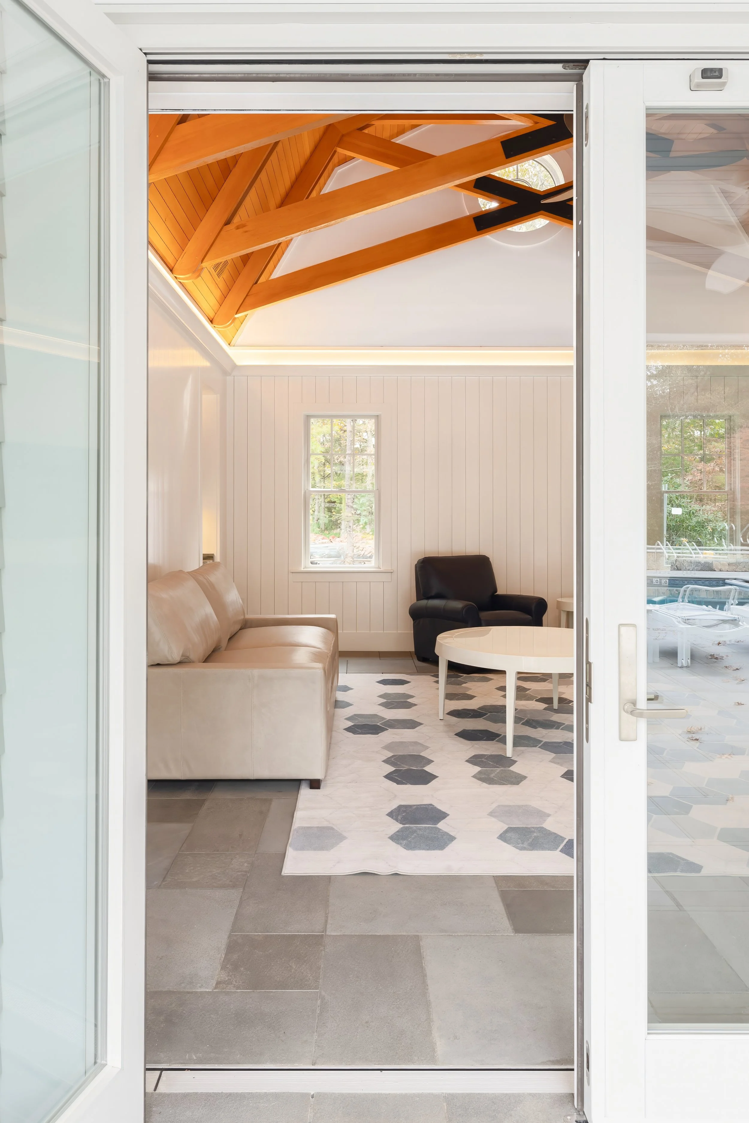 Sliding glass doors open to a custom Cape Cod pool house with exposed wood trusses, vaulted ceilings, and direct access to the outdoor living and pool area.