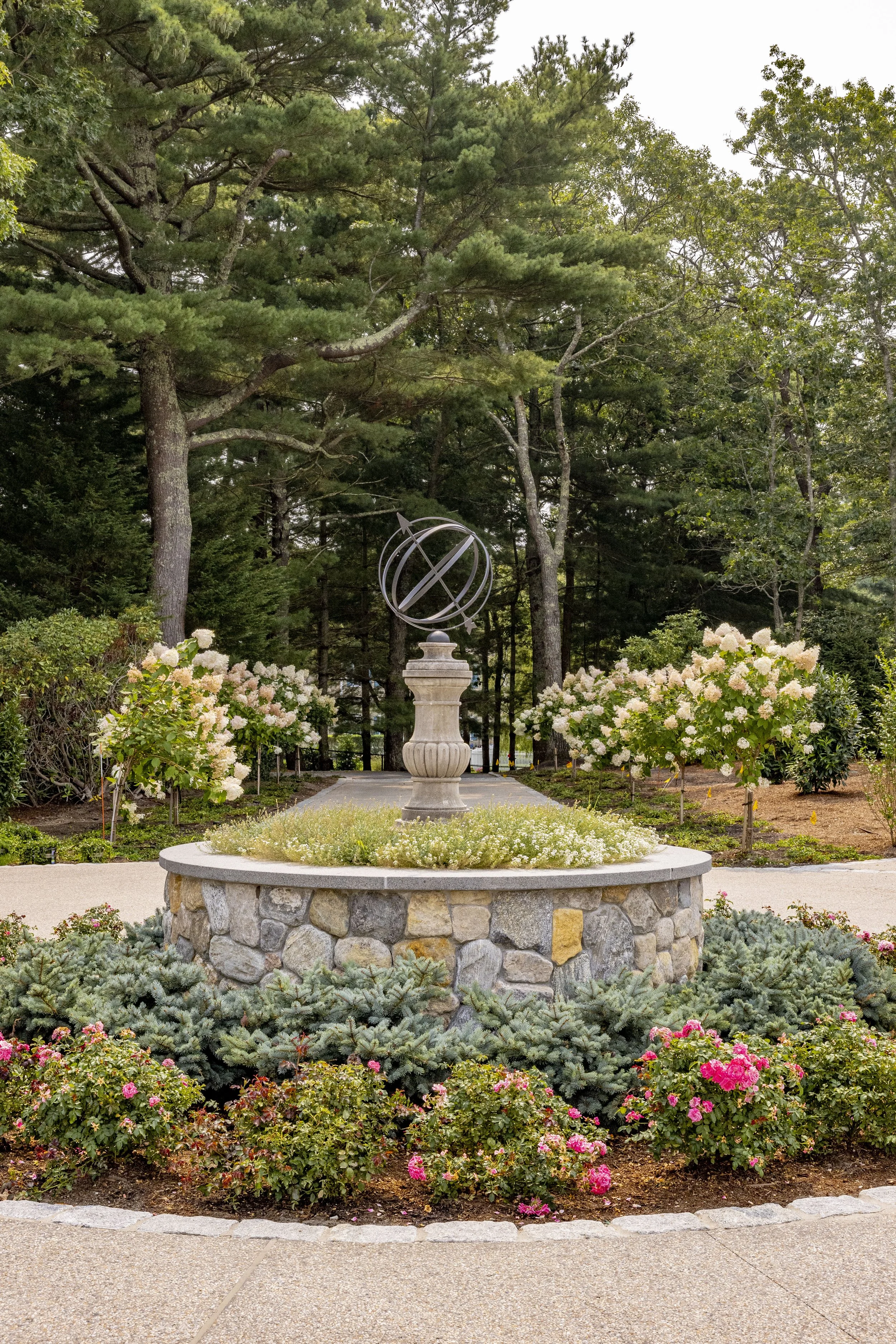 Circular stone fountain with garden flowers and sculpture framed by tall trees in a Cape Cod landscape.
