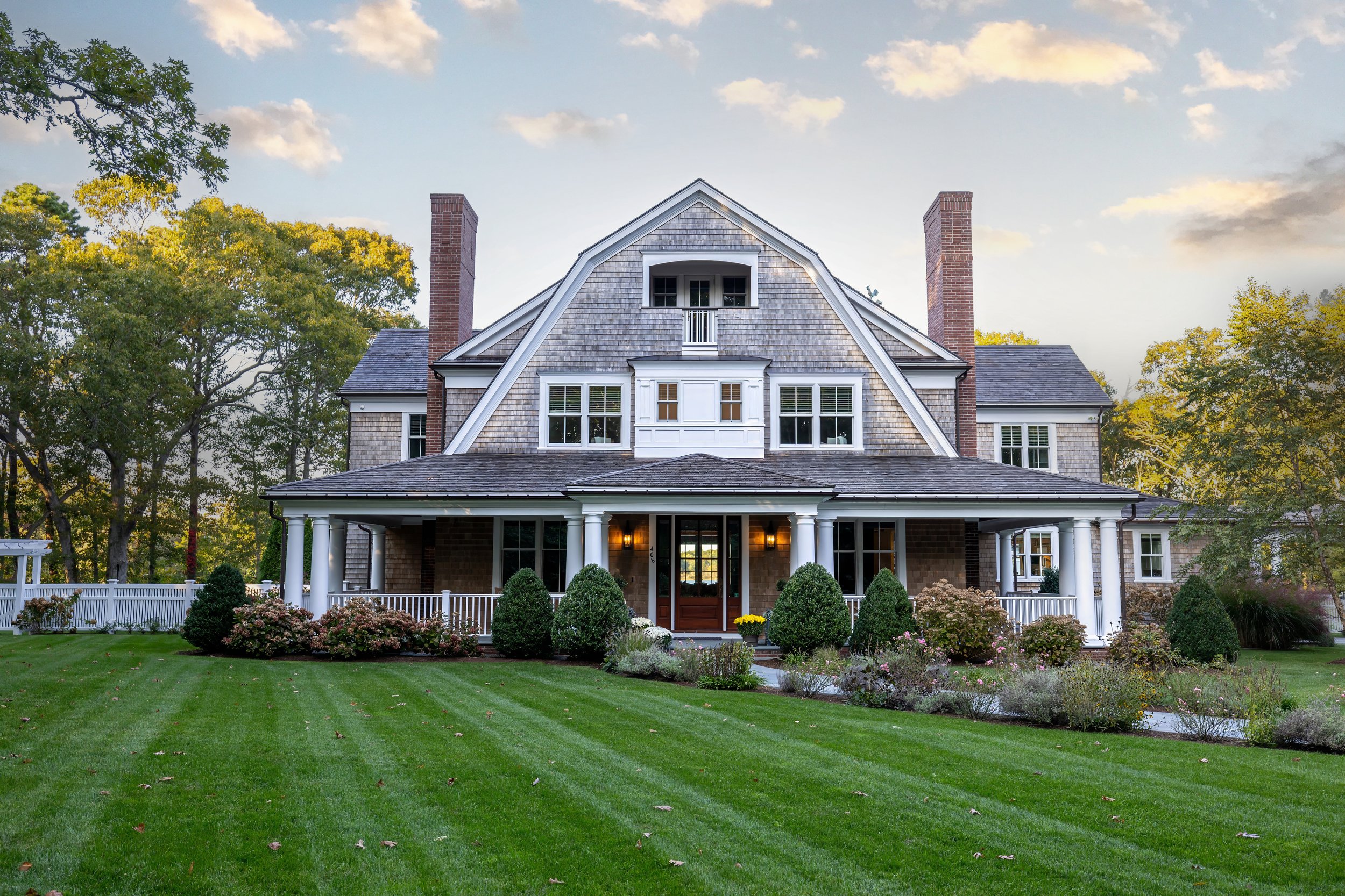 Grand Cape Cod home with gambrel roof, white columns, brick chimneys, and landscaped front lawn.