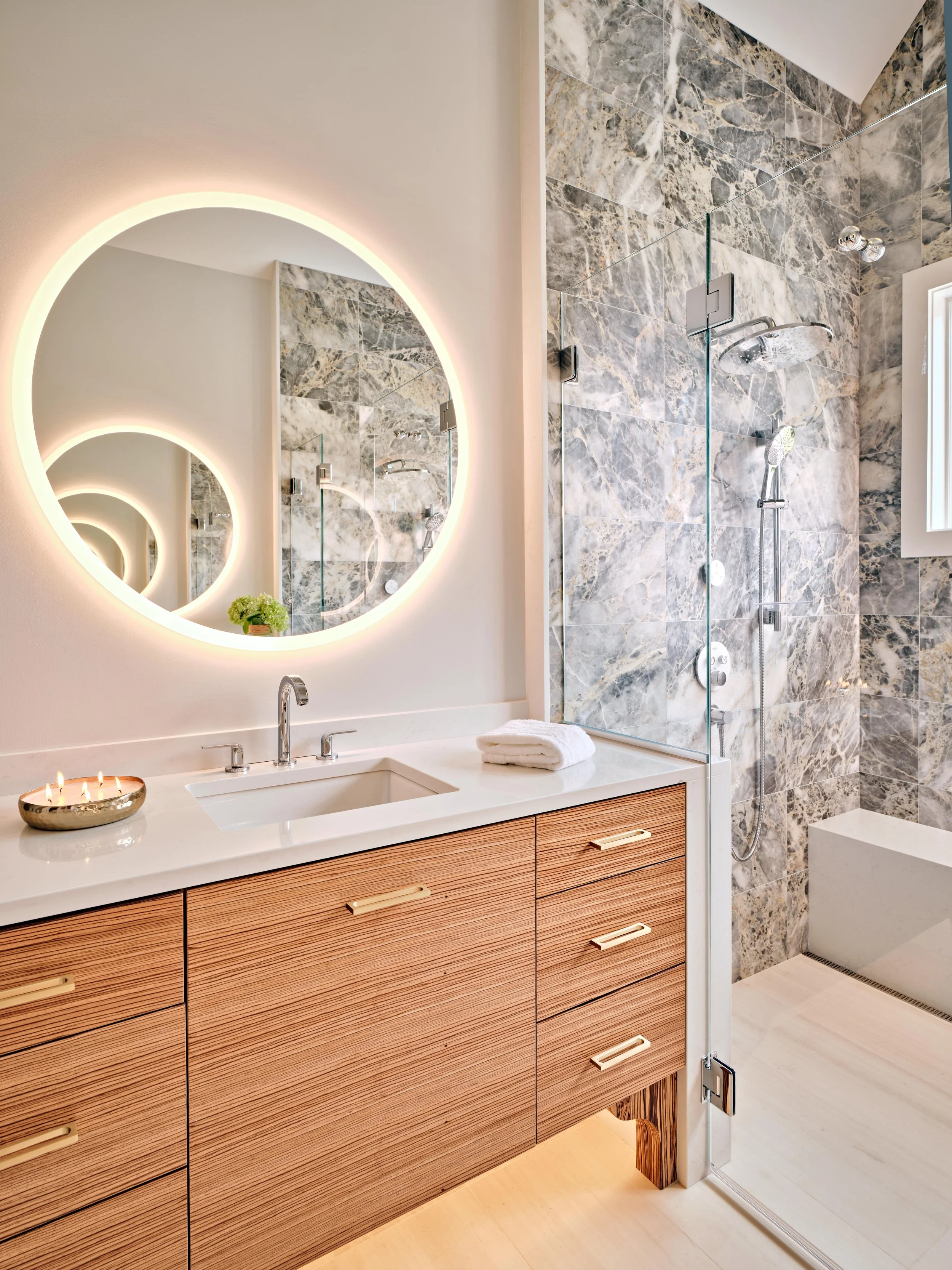 Spa-like bathroom with illuminated round mirror, wood vanity, and marble shower in a high-end Cape Cod renovation.