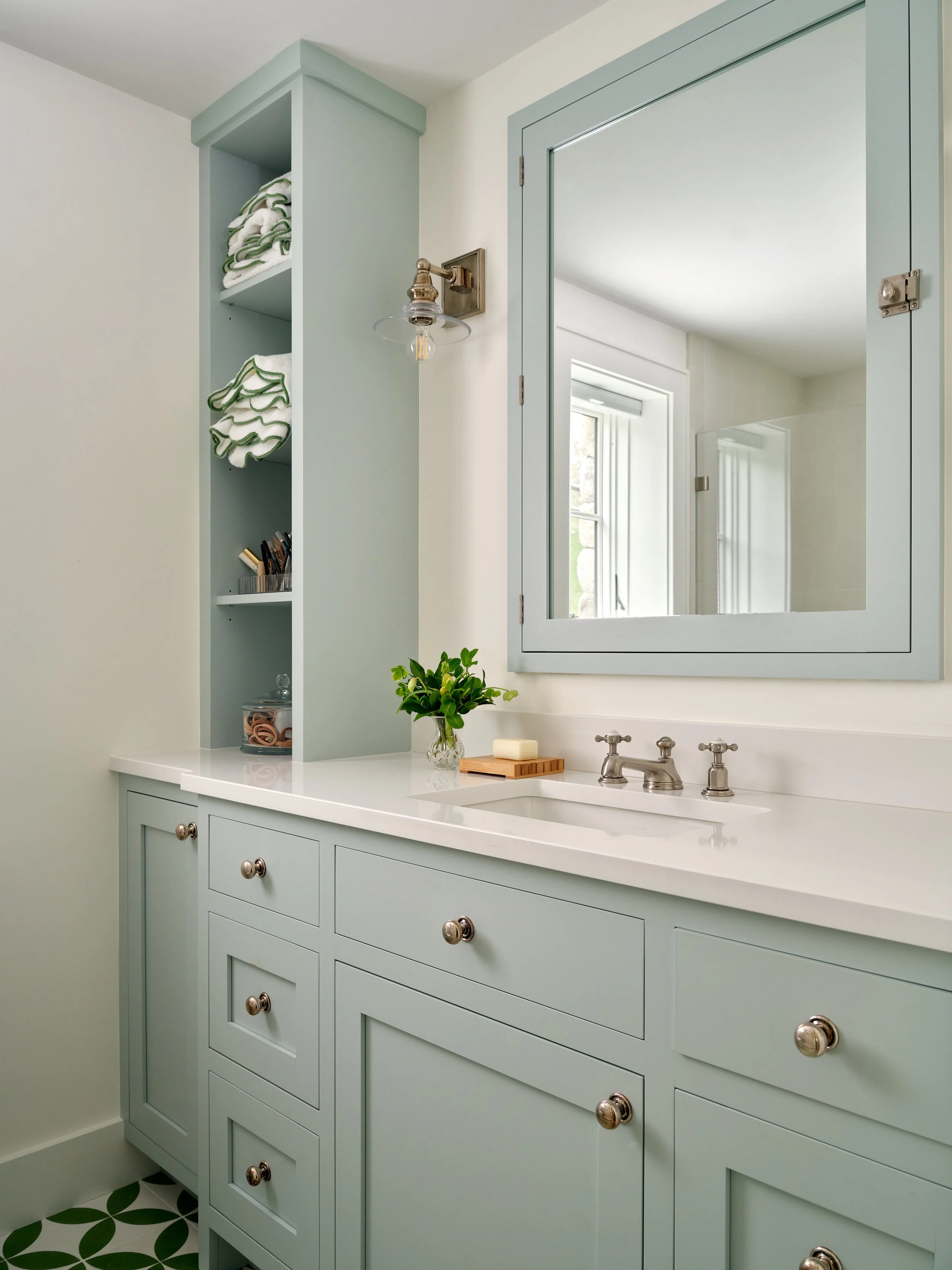 Blue bathroom vanity with open shelving, framed mirror, polished hardware, and patterned tile flooring.