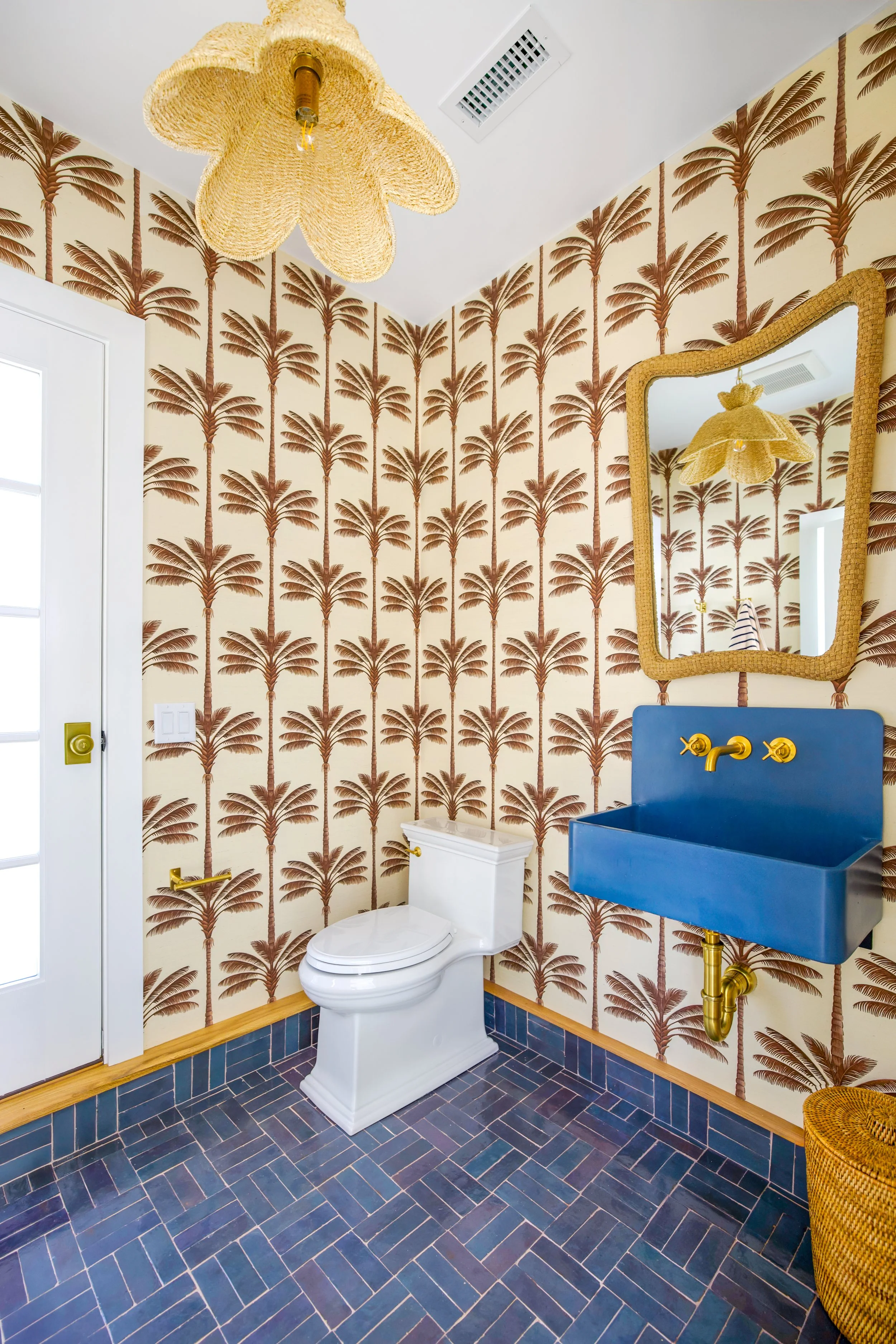 Palm wallpaper, blue sink, and herringbone tile bring playful coastal powder room design to this space.