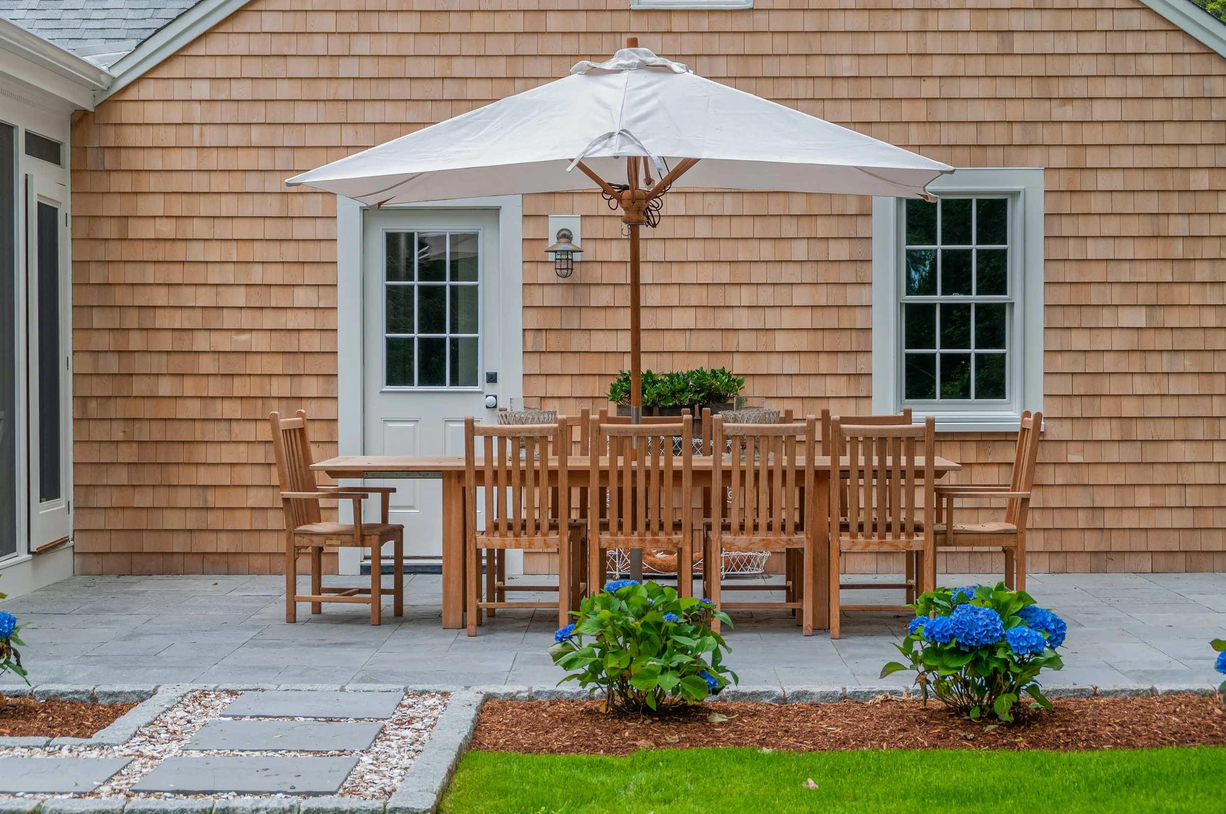 Patio dining area beside a cedar-shingled Cape Cod home with umbrella, blue hydrangeas, and stone pavers.