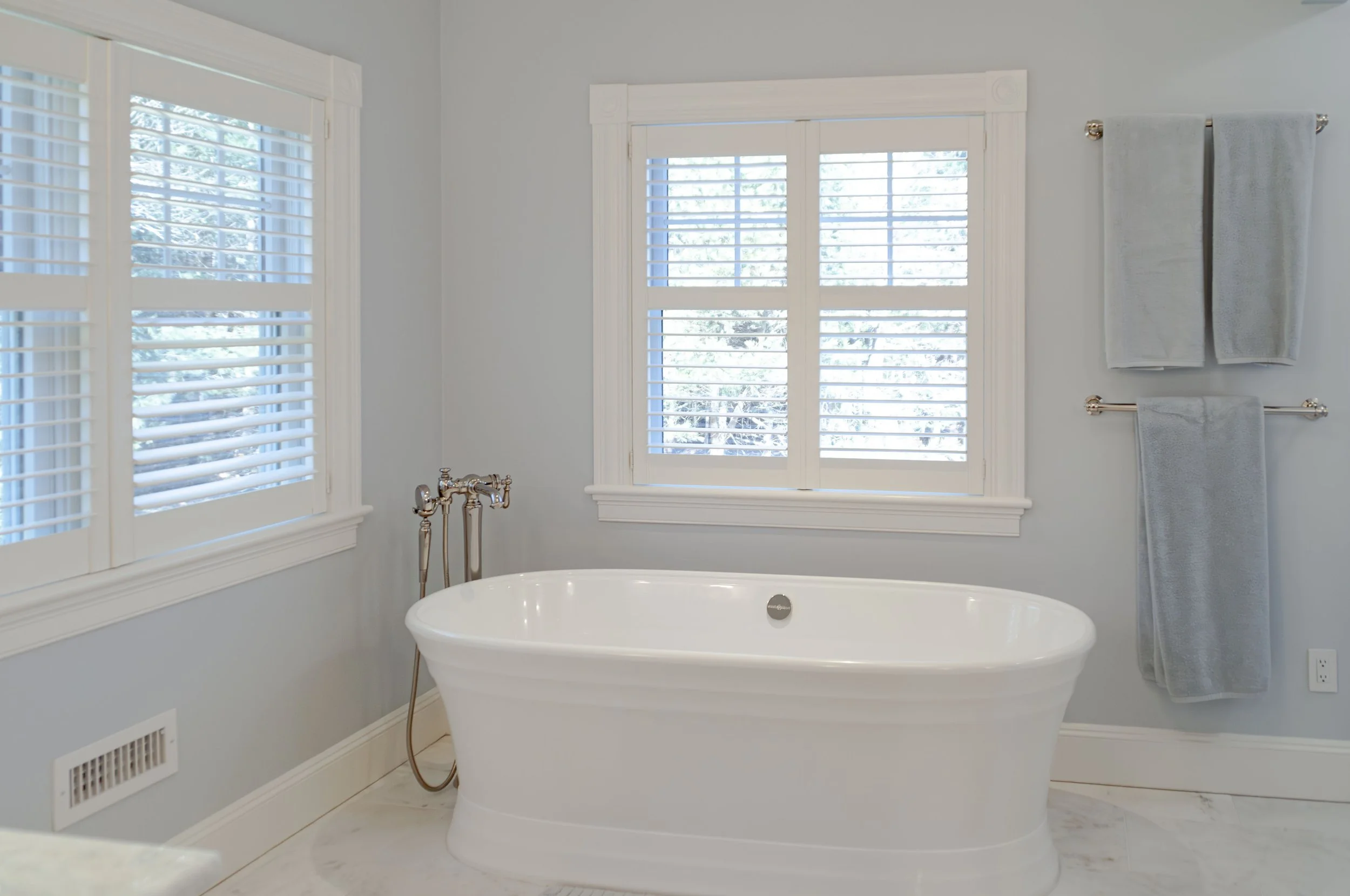 Cape Cod bathroom with freestanding tub, shuttered windows, marble floor, and soft blue walls.
