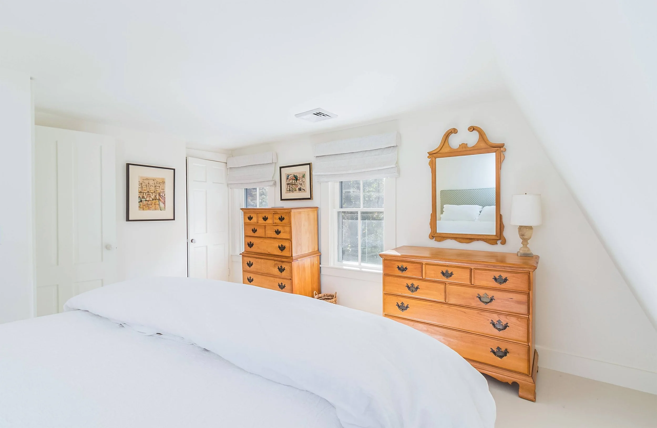 airy white bedroom with wood dressers, soft window shades, and classical coastal home detailing. 
