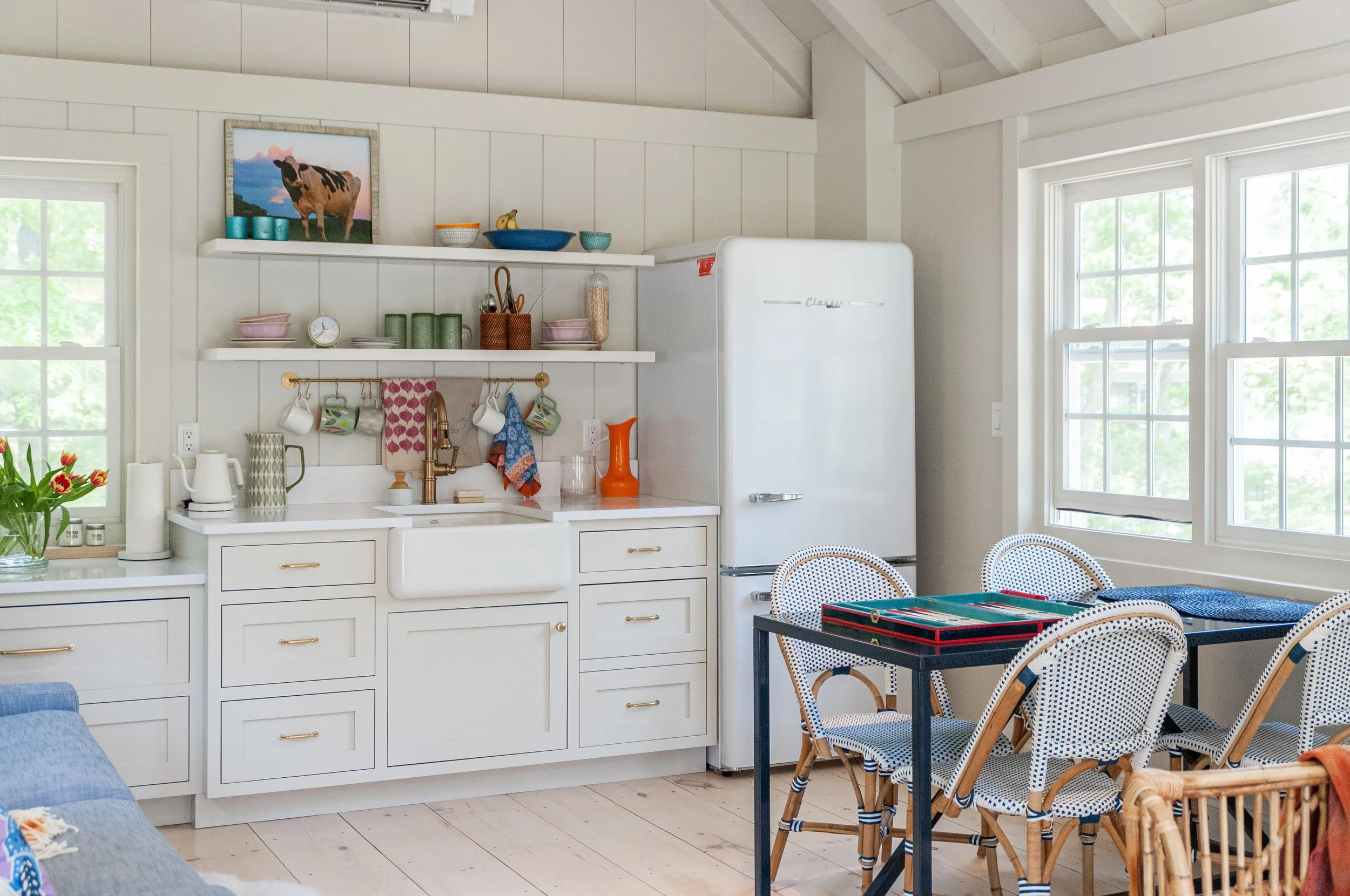 A cheerful custom coastal home kitchenette features open shelving, farmhouse sink, vintage-style refrigerator, and sunny dining area.