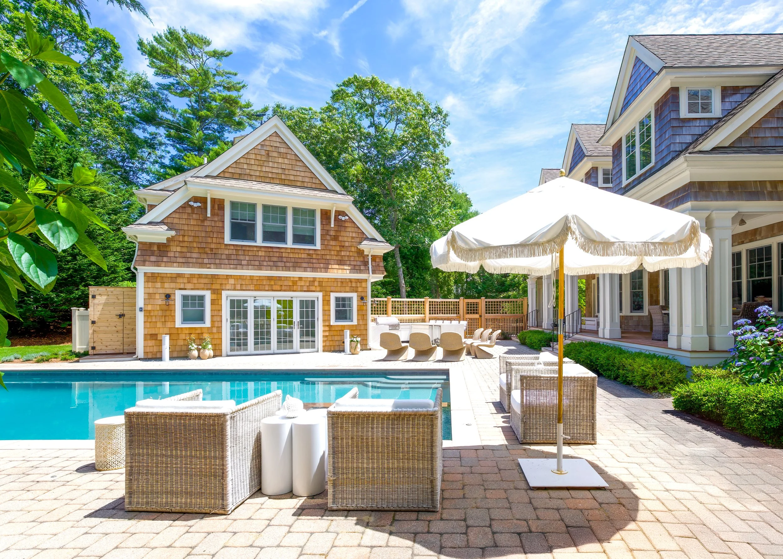 Shingled pool house, wicker seating, umbrellas, and blue pool define this Cape Cod outdoor living space.
