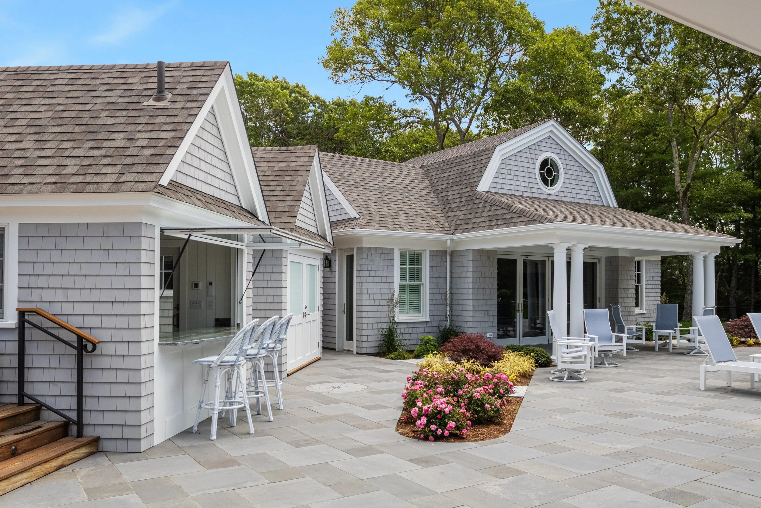 Shingle-style Cape Cod pool house with bar seating, covered patio, lounge chairs, and garden plantings.
