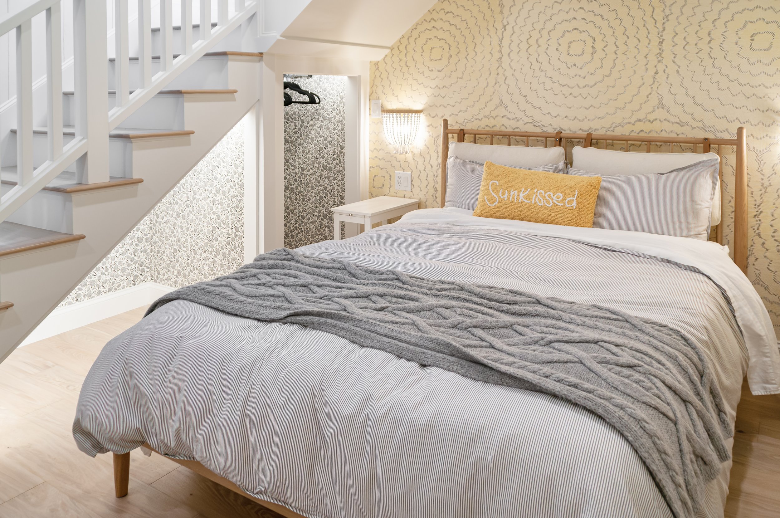 Cozy bedroom tucked beneath a white staircase, featuring patterned wallpaper, wood floors, and layered bedding.