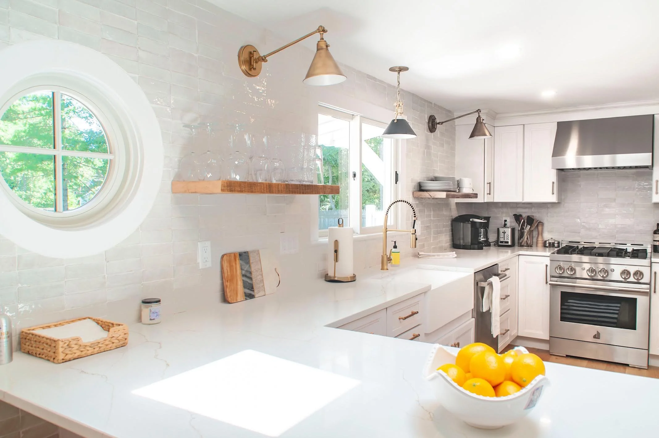 Glossy tile, round window, brass lighting, and stainless appliances shape this bright custom Cape Cod kitchen.