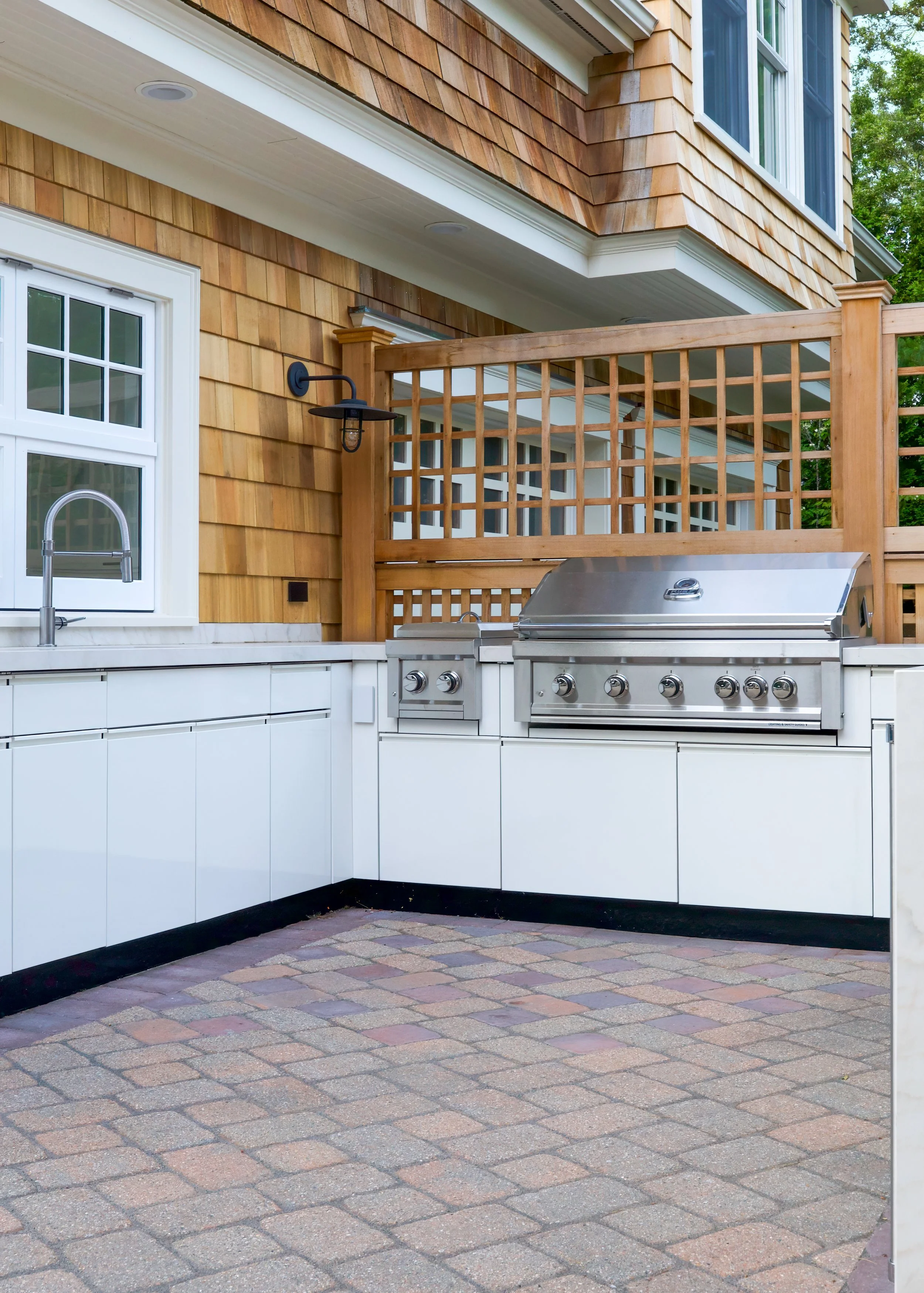 Built-in grill, sink, and cedar lattice define this Cape Cod outdoor kitchen design beside shingled siding.