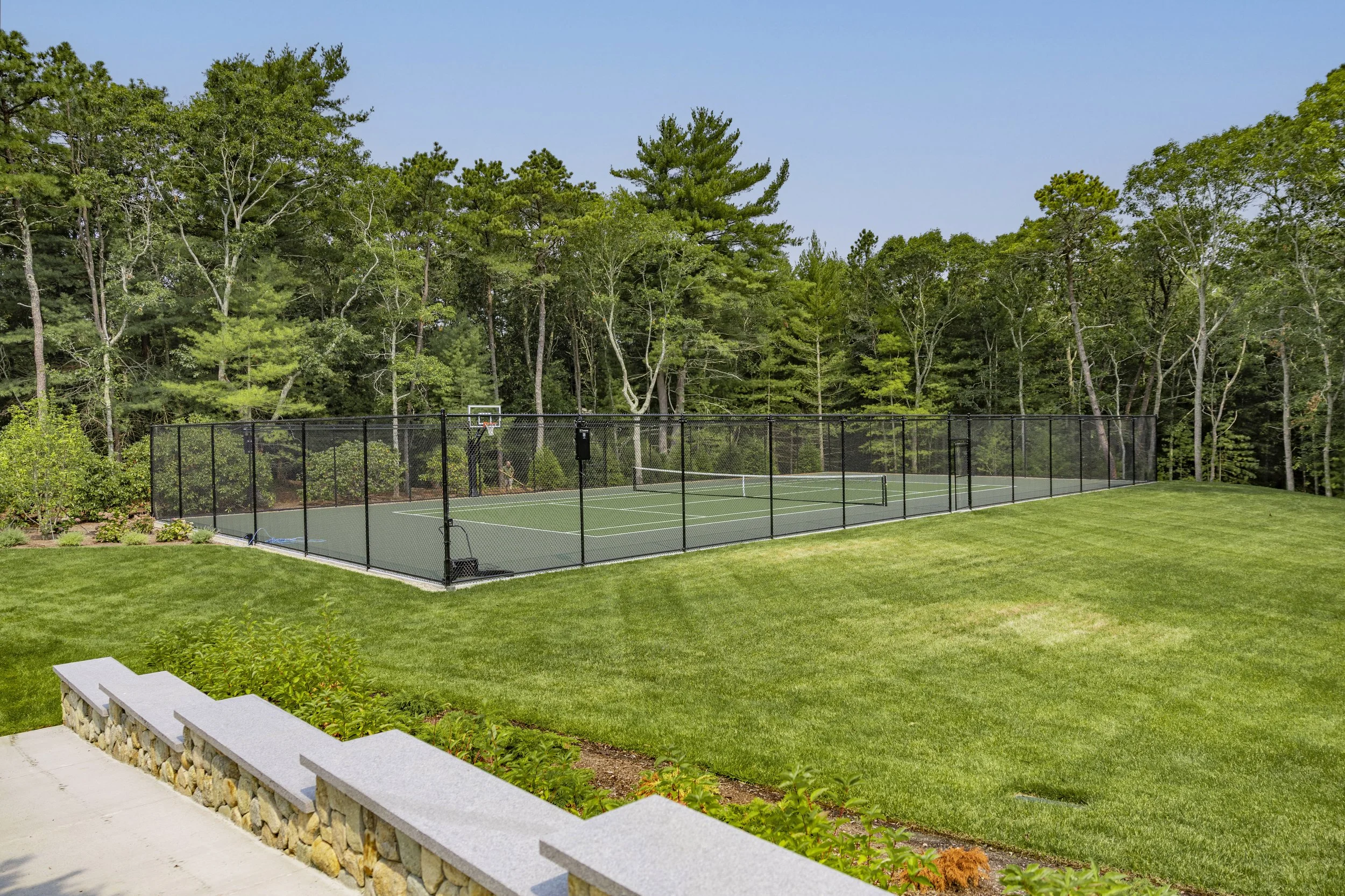 Cape Cod property with fenced tennis, pickleball, and basketball court beside manicured lawn and woods.