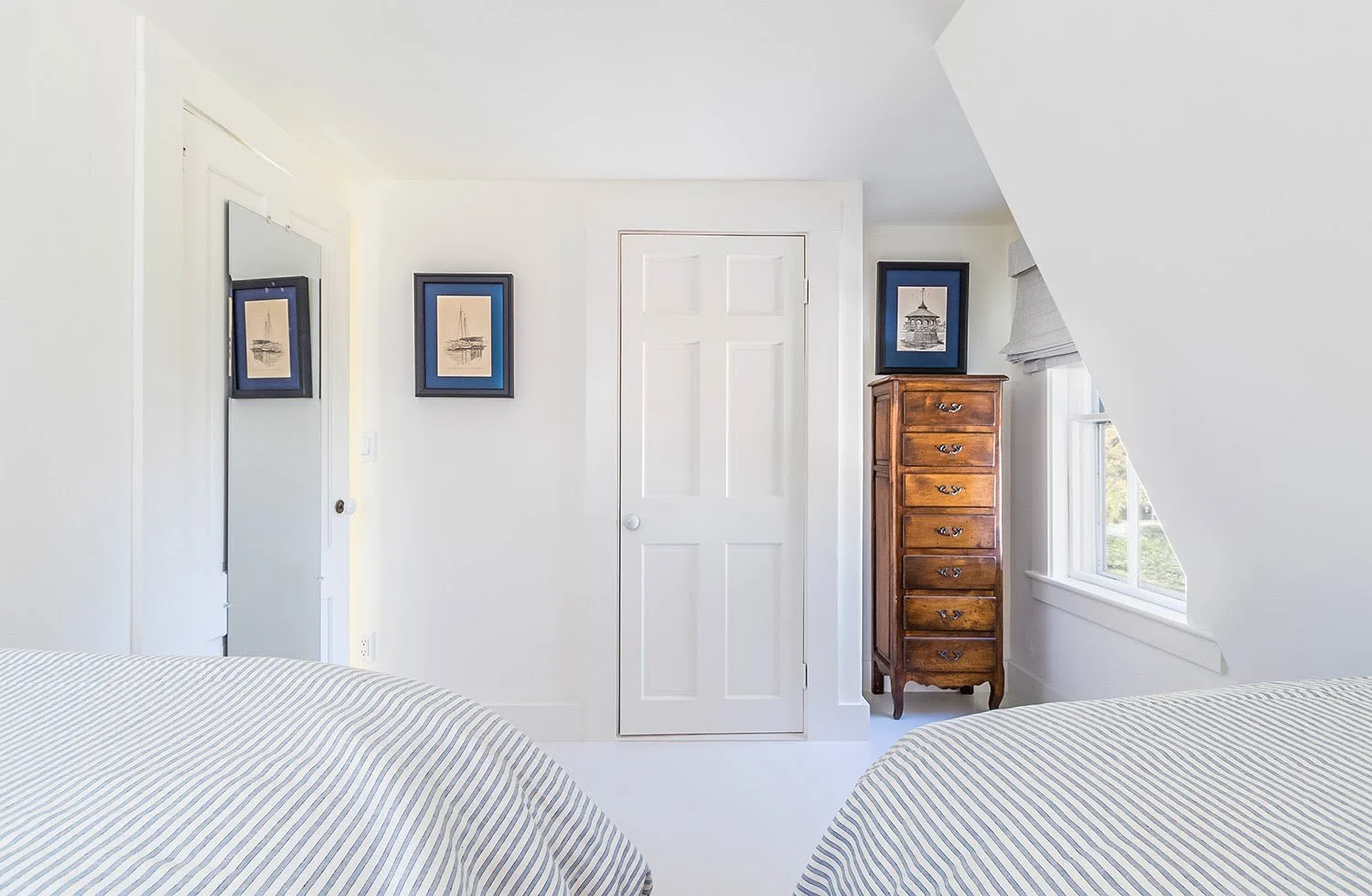 Striped twin beds, angled ceilings, and framed art create a bright Cape Cod cottage guest room.