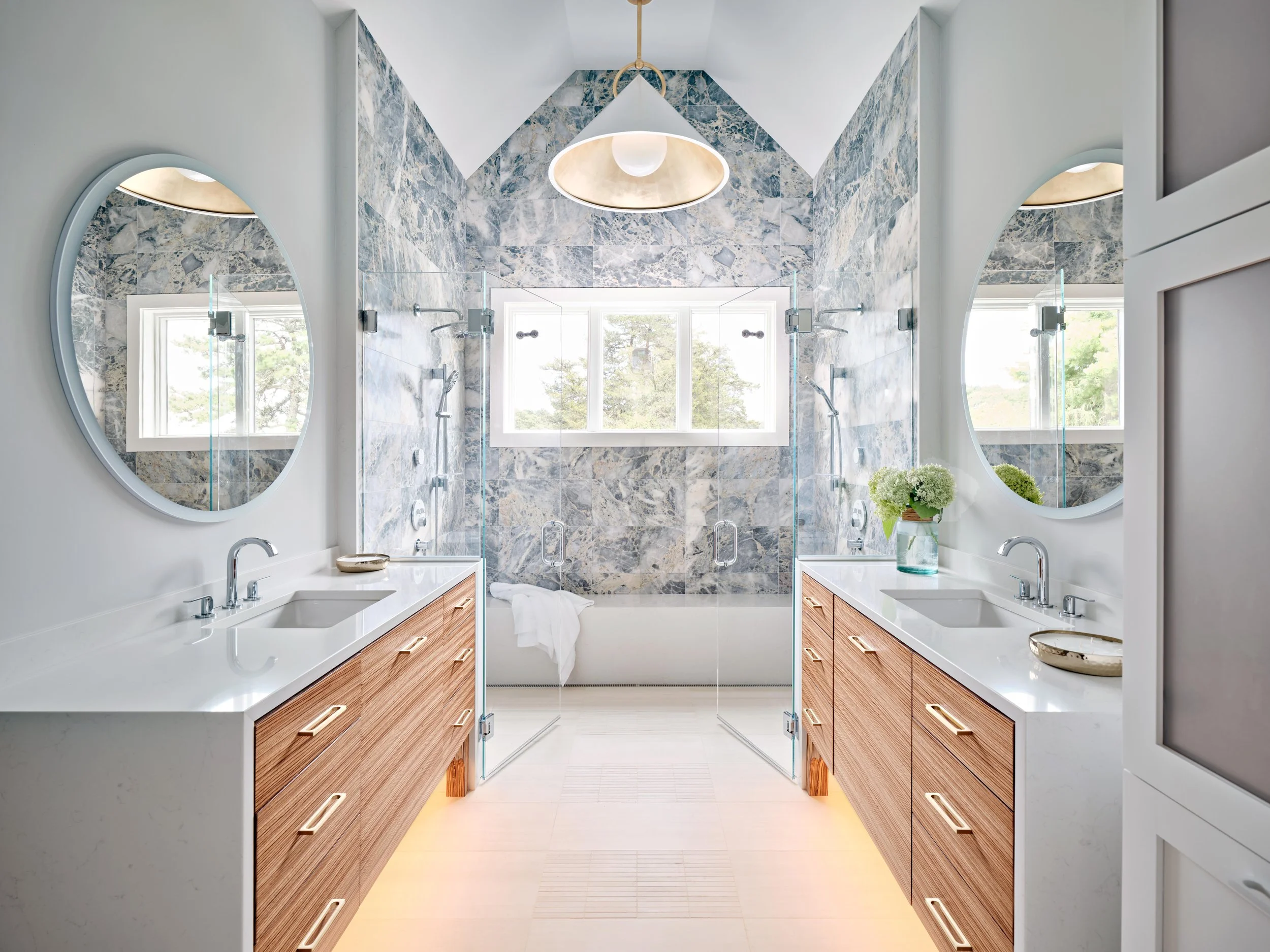 a custom coastal home bathroom features twin vanities, round mirrors,glass shower doors, and marble-style tile. 