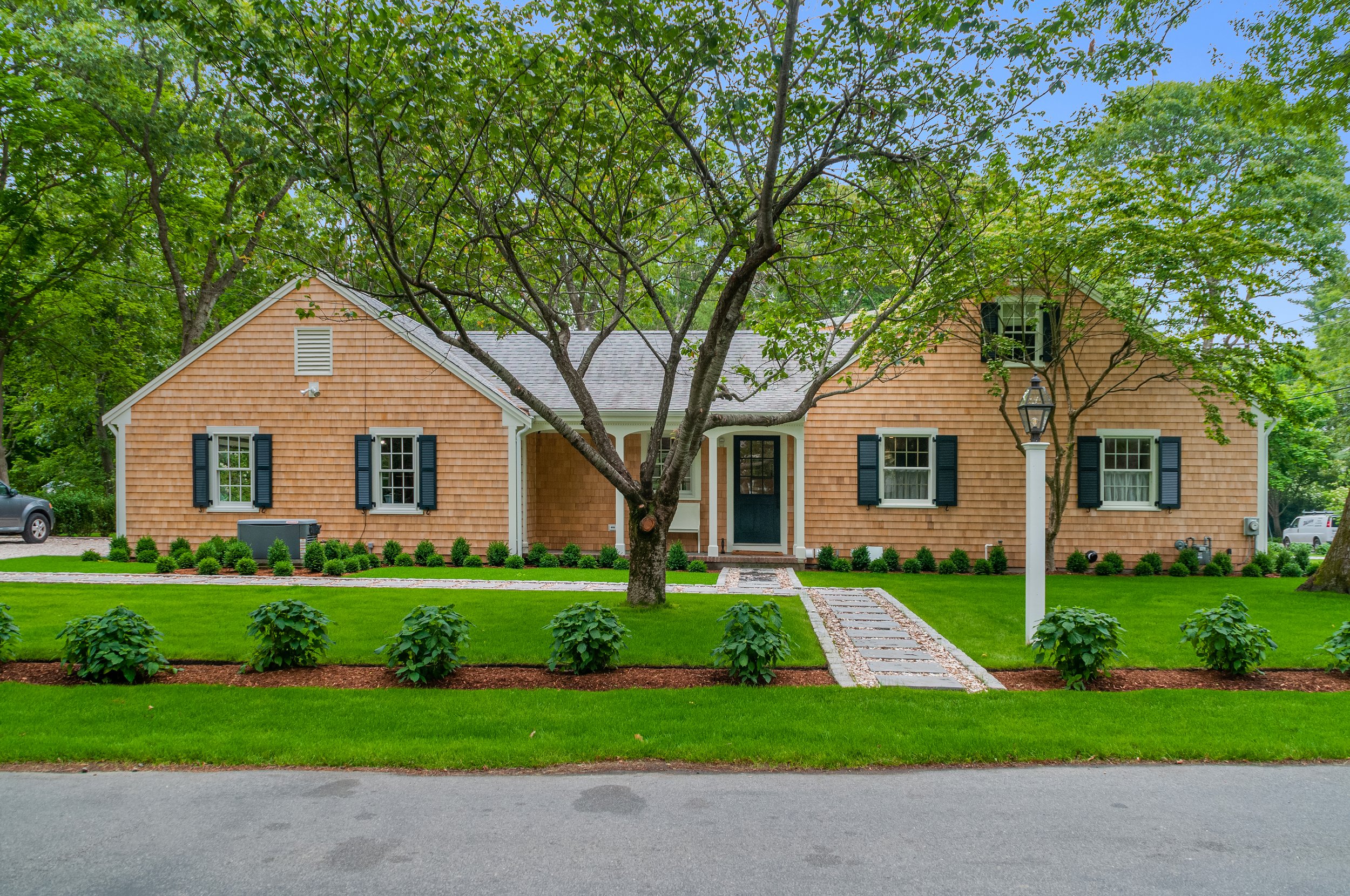 Cedar-shingled Cape Cod residence with black shutters, stone walkway, mature trees, and tailored coastal landscaping.