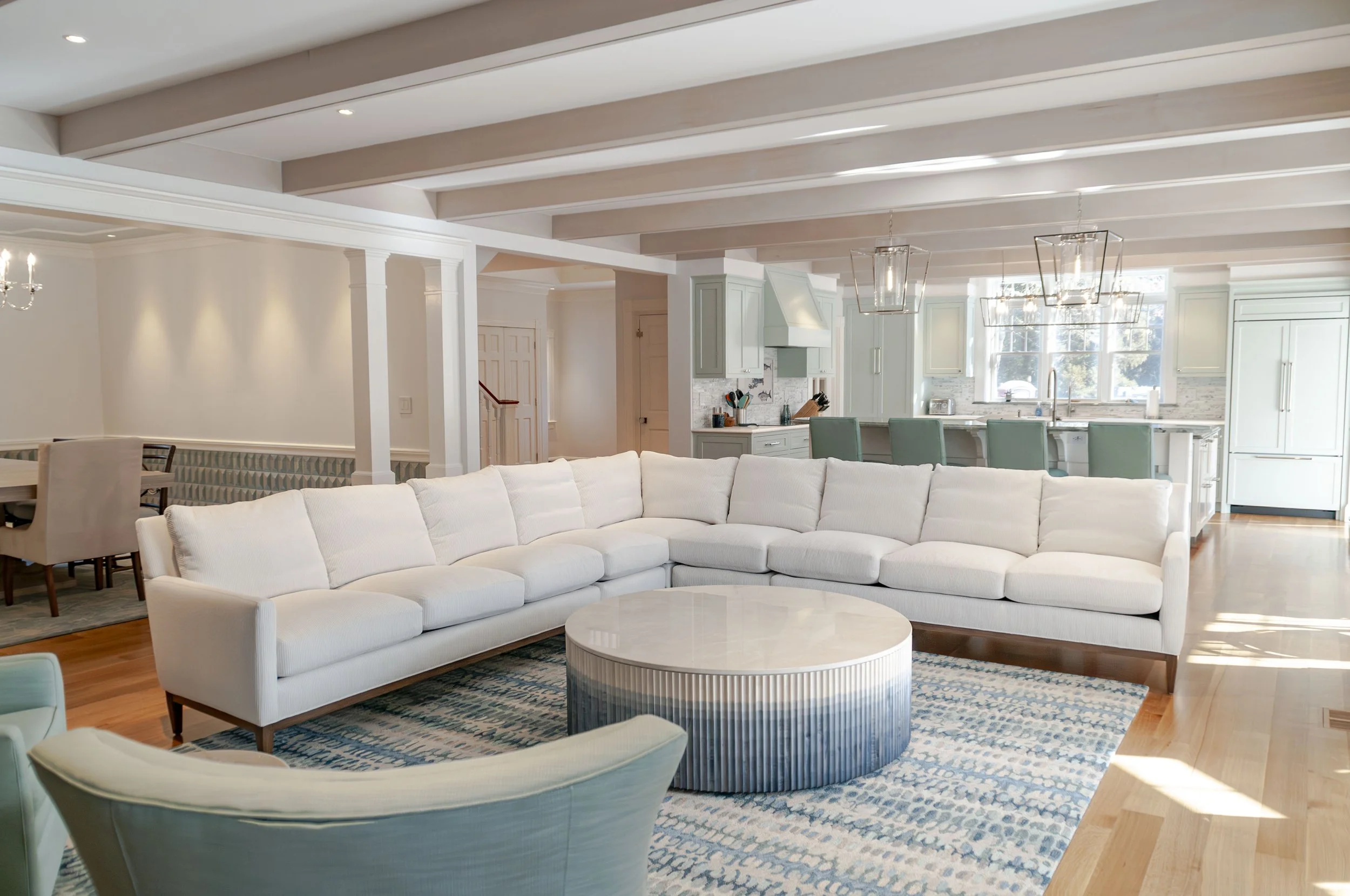 Open Cape Cod living room with white sectional, blue patterned rug, beamed ceiling, and kitchen beyond.
