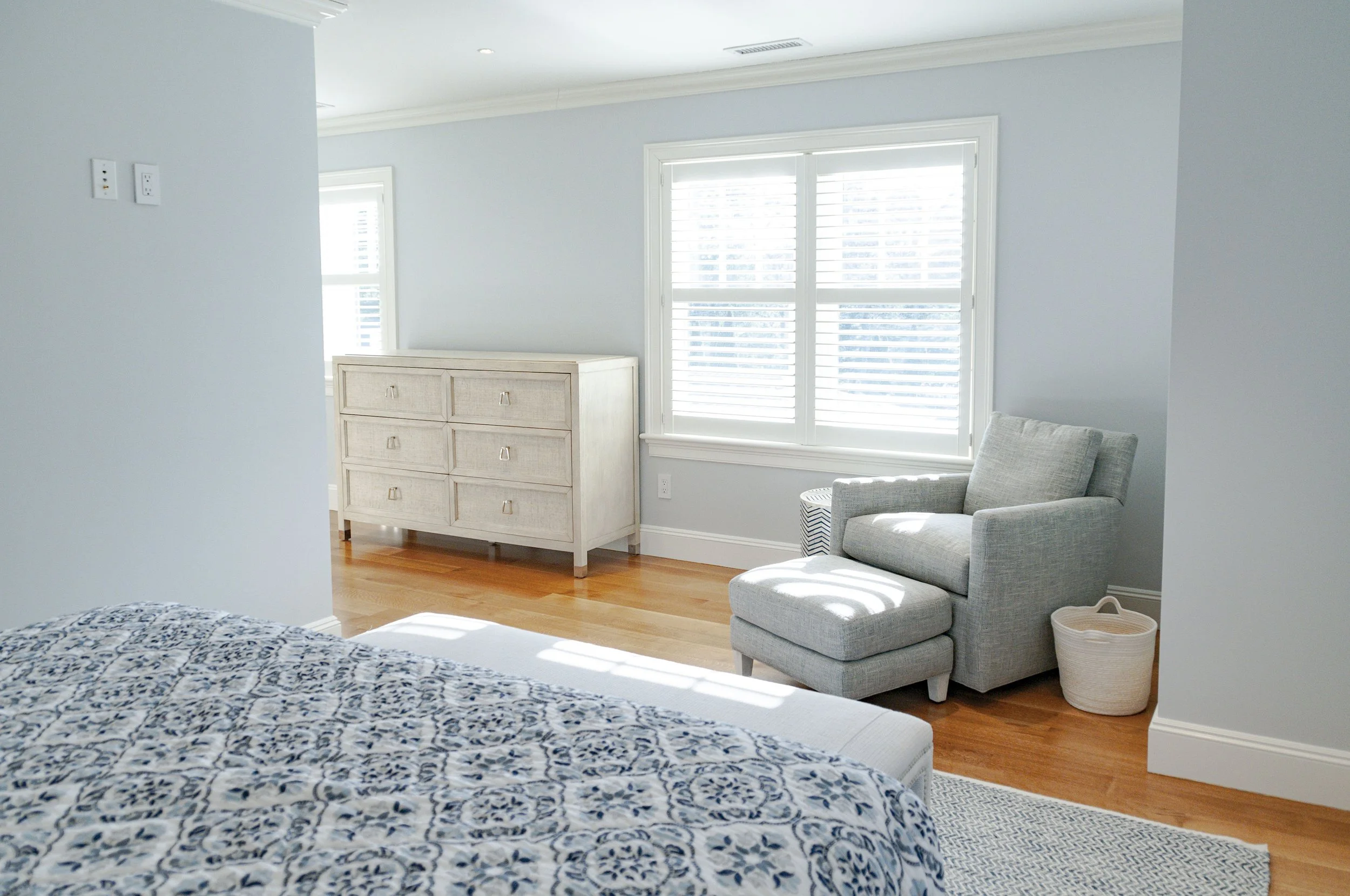 Bright primary bedroom in a Cape Cod custom home with patterned bedding, lounge chair, and shuttered windows.