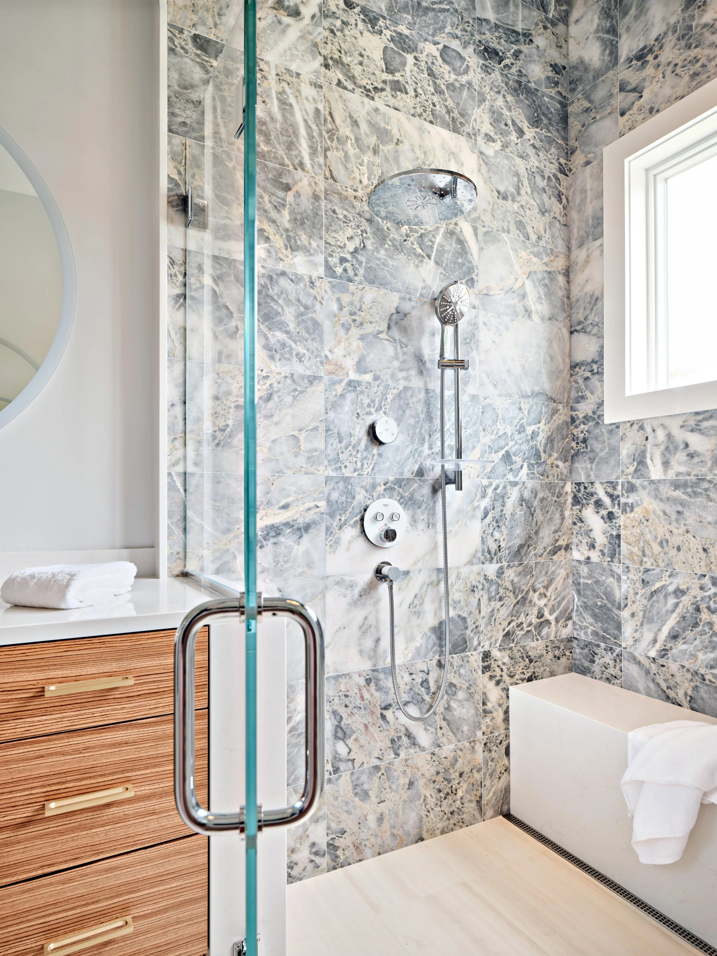 This high-end Cape Cod renovation features a glass shower, marble tile, wood vanity, and polished chrome fixtures.