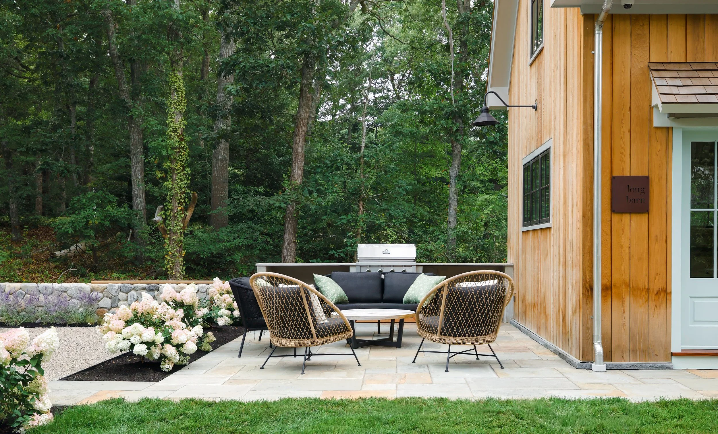 Custom coastal home patio with woven chairs, outdoor grill, stone wall, and wooded Cape Cod setting.