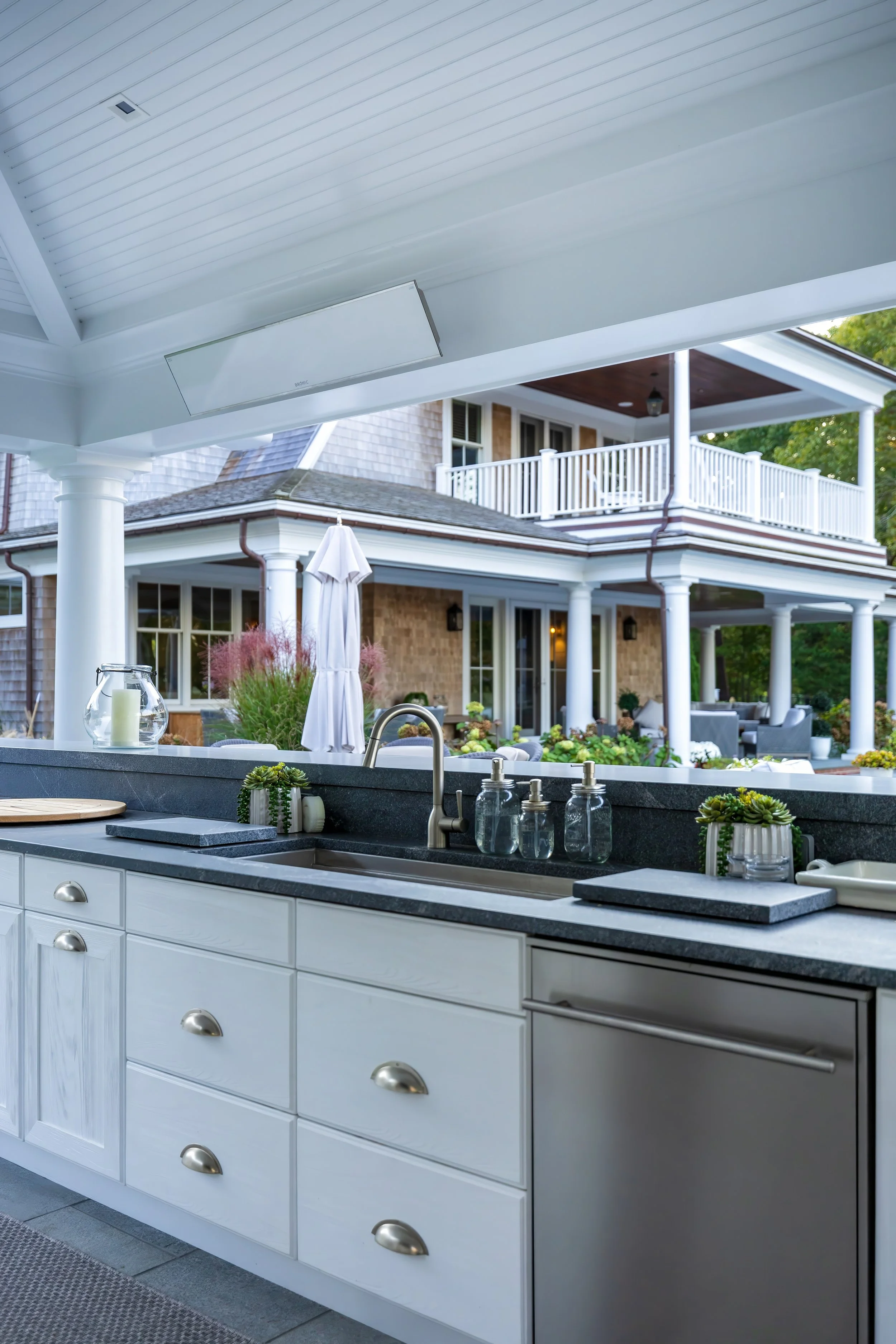 Outdoor kitchen sink and cabinetry with black countertop, white drawers, and a luxury Cape Cod home beyond.