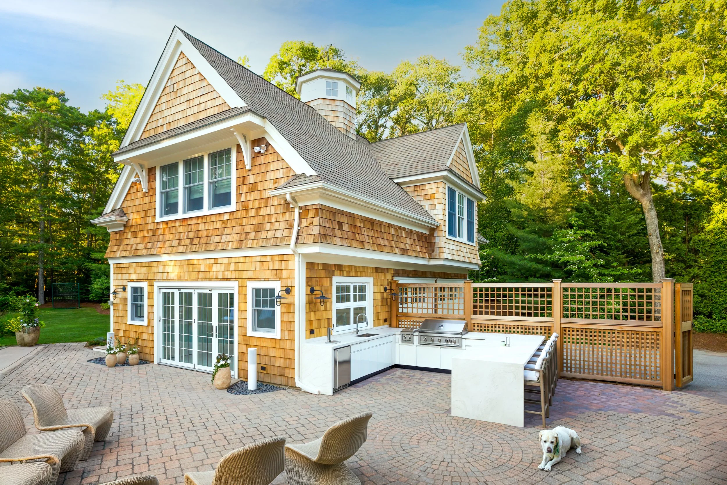Luxury Osterville carriage house featuring a custom outdoor kitchen, cedar shingles, spacious patio, outdoor entertaining area, and Cape Cod landscape design.