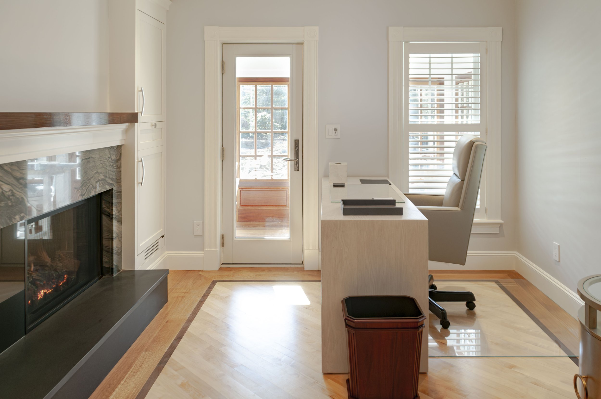 Home office with fireplace, glass door, herringbone floor, and bright windowed workspace.