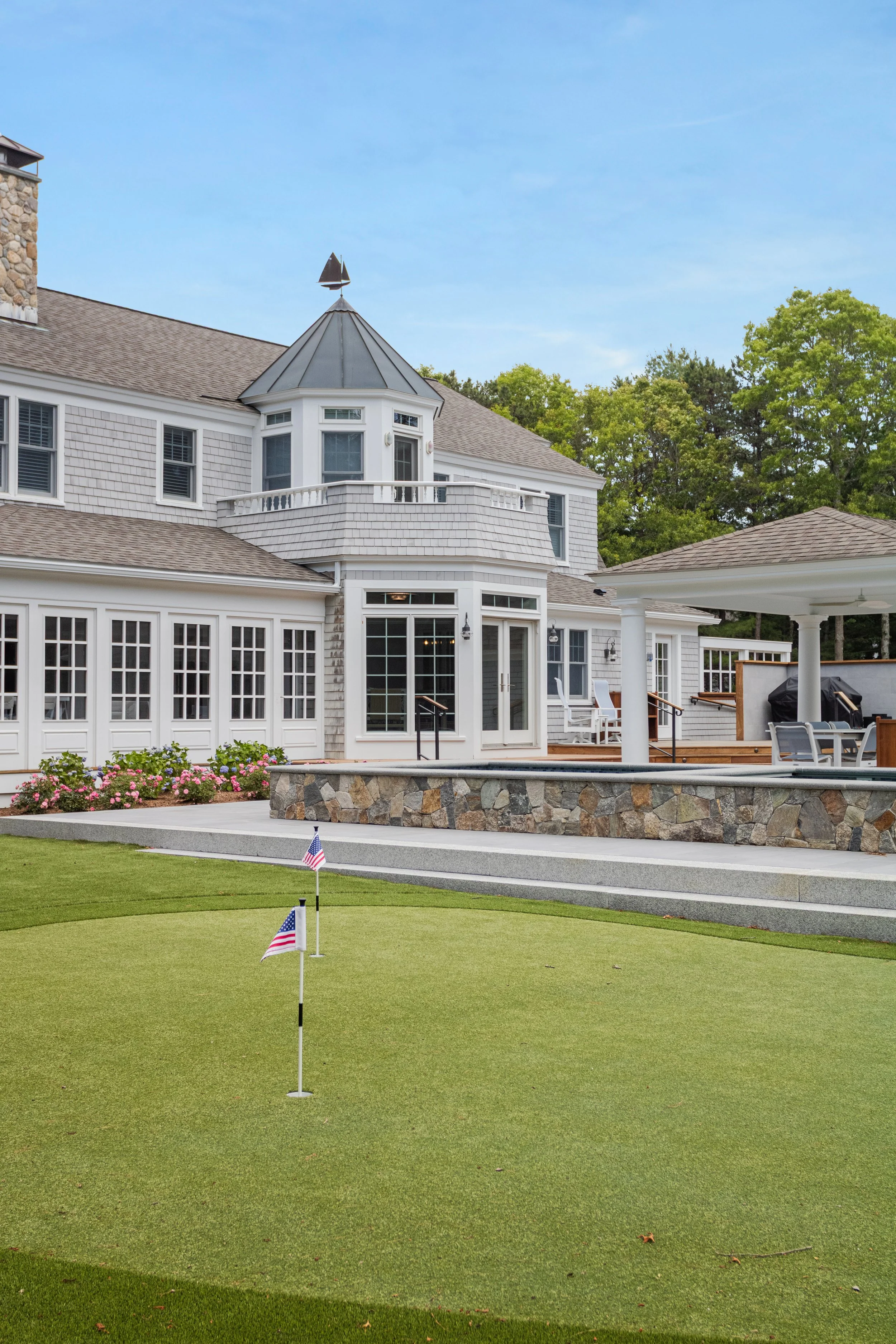 Resort-style backyard featuring a custom putting green, covered outdoor pavilion, stone landscaping, and a waterfront-inspired Cape Cod custom home.