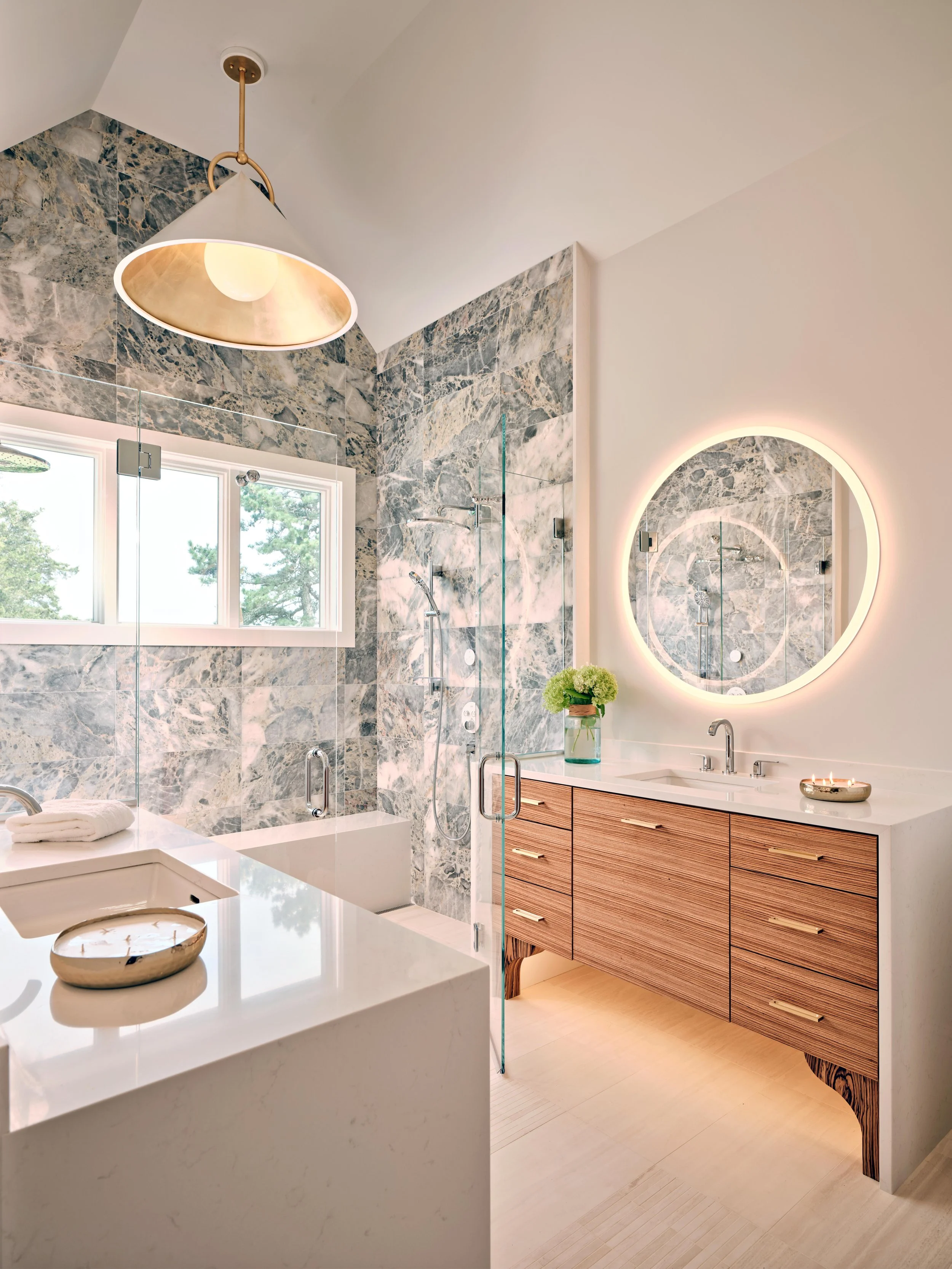 A bright bath in this high-end Cape Cod renovation showcases marble tile, glass shower, wood vanity, and illuminated mirror.